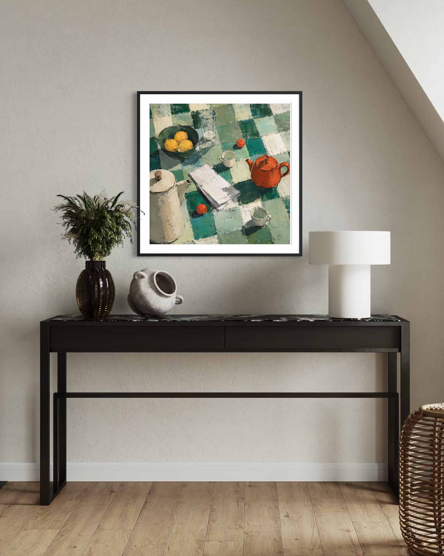 A framed art print featuring a still life painting with a green and white checkered pattern, yellow lemons, and a red teapot, displayed on a beige wall above a black console table with decorative items.