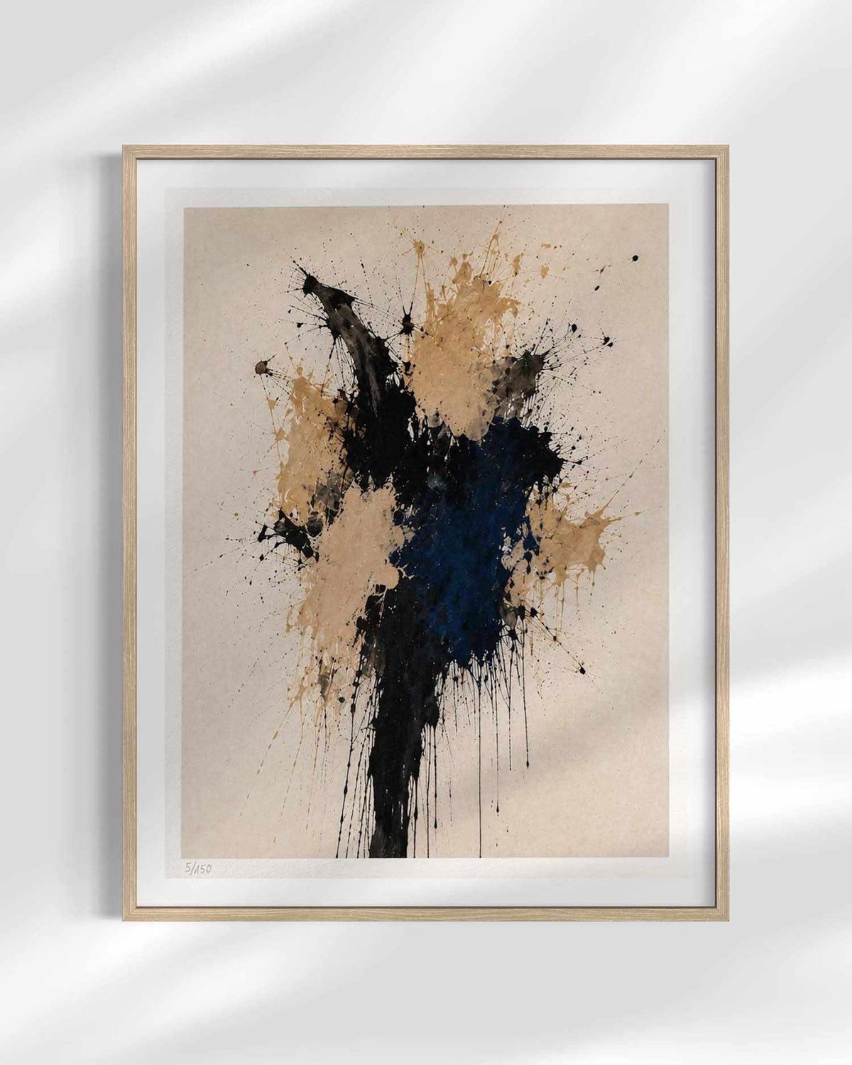 Framed abstract art print featuring a textured paint splatter design in black, navy blue, and tan on a warm beige background, housed in a light natural wood frame with a white mat.