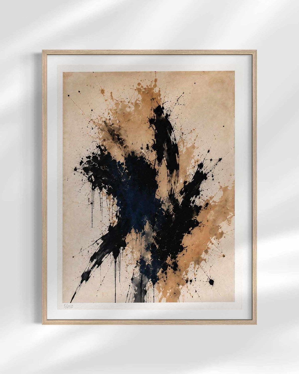 Framed abstract wall art print with a light wood frame and white mat, featuring black and dark blue paint splatters over a warm beige textured background, shown hanging on a white wall with soft shadow overlays.