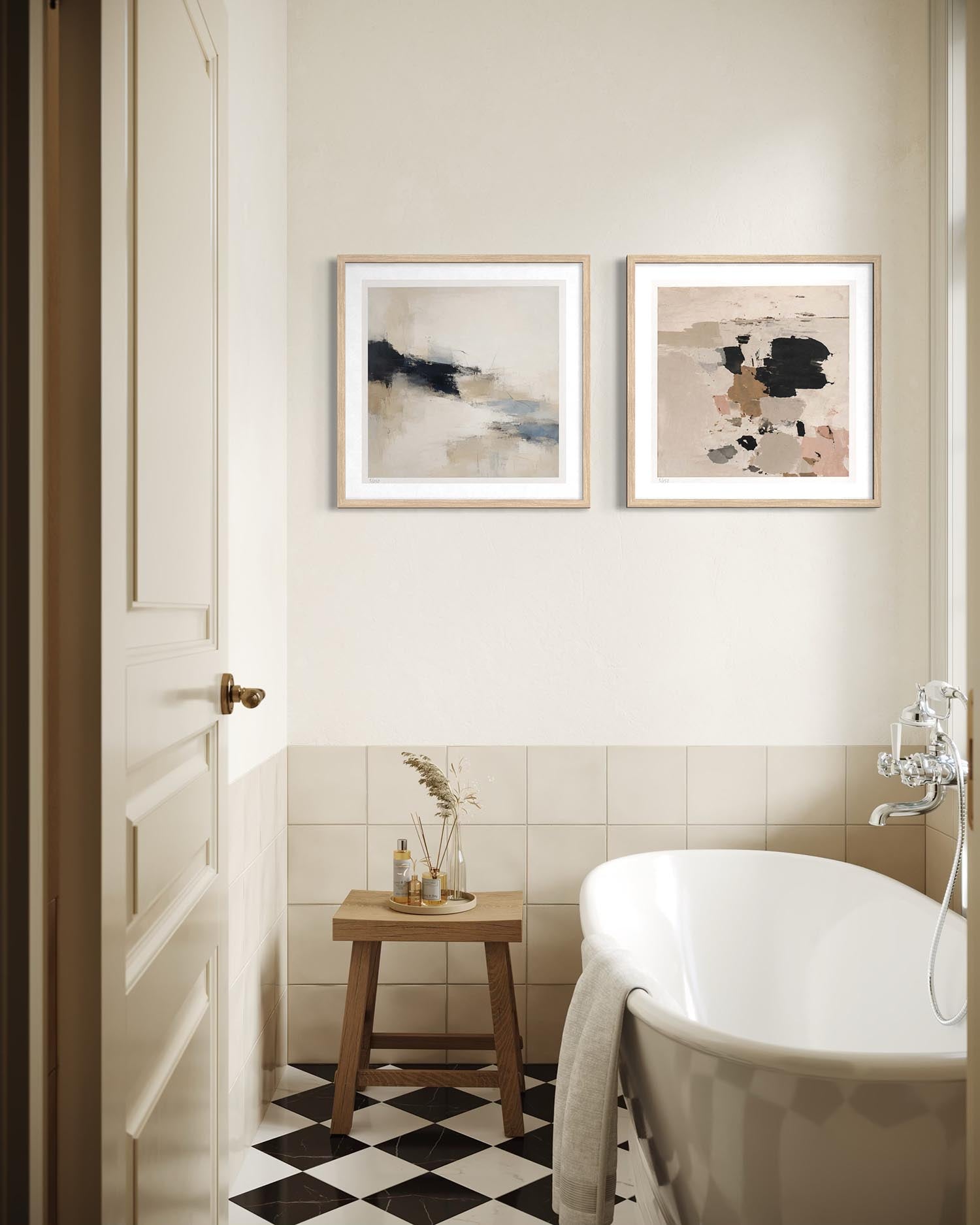 Two framed abstract art prints hang on a cream-colored bathroom wall above a white bathtub and a small wooden stool.