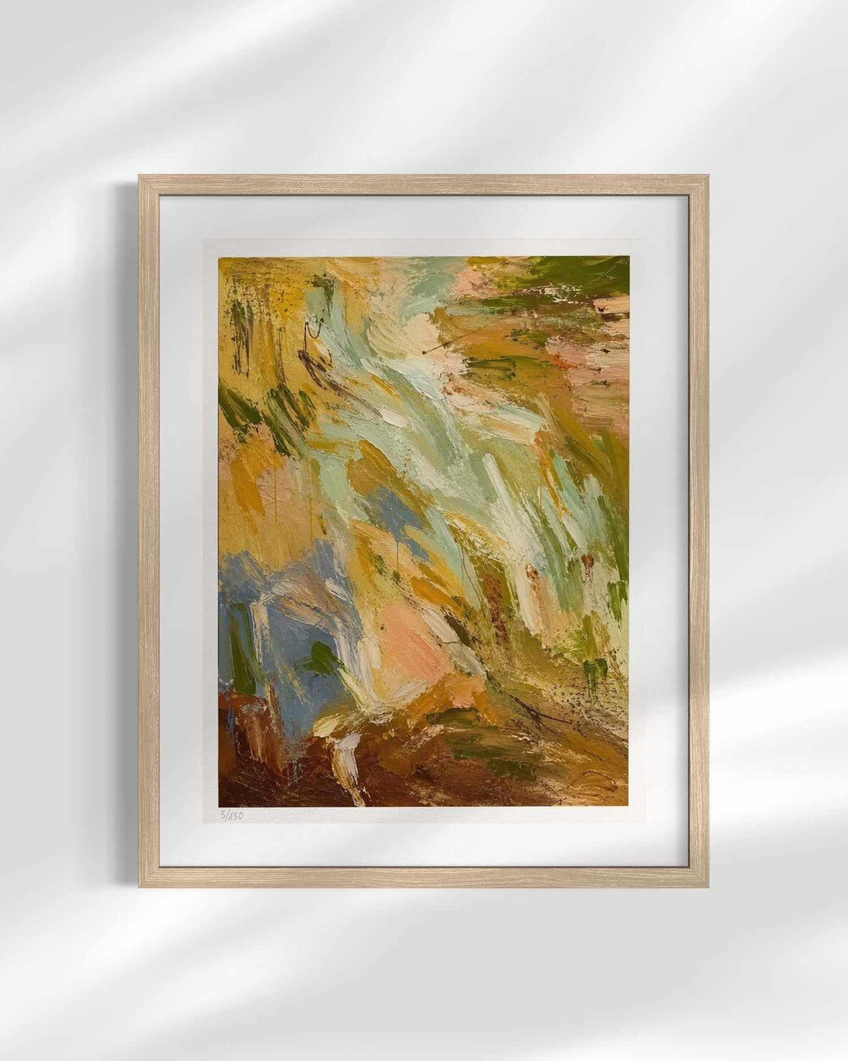 A framed abstract painting with expressive brushstrokes in earthy tones of brown, gold, green, blue, and cream, displayed in a light wood frame with a white mat against a softly lit wall.