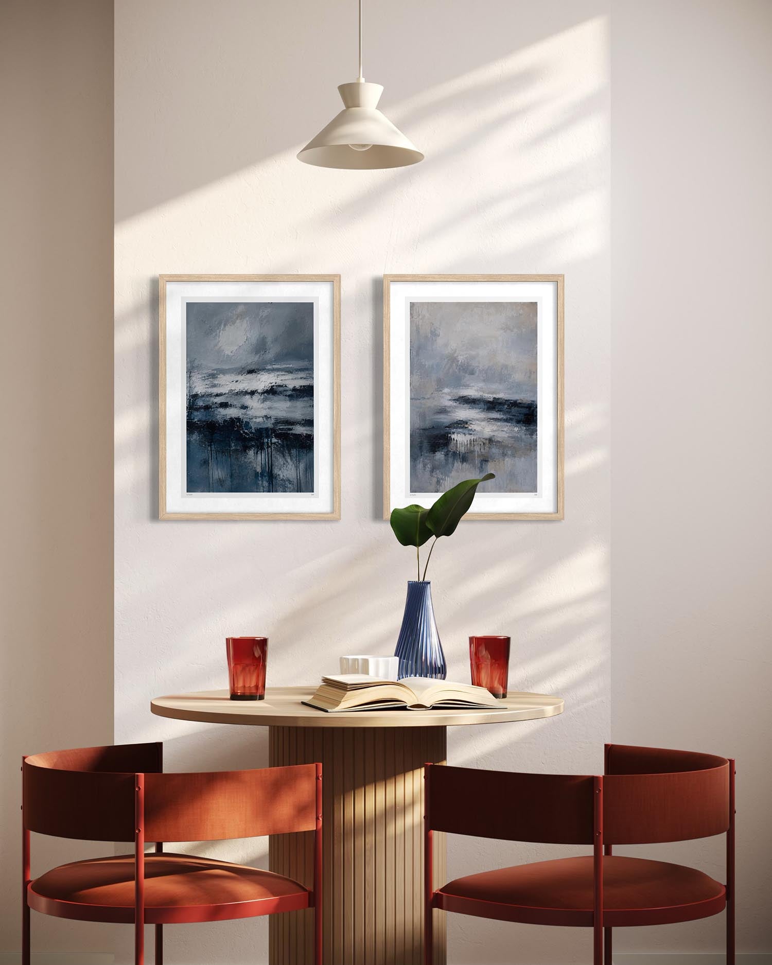 A styled dining room corner with two framed abstract blue and gray wall art prints above a round light wood table, flanked by two rust-red chairs. A white pendant light hangs overhead, and a blue vase with green leaves sits on the table.