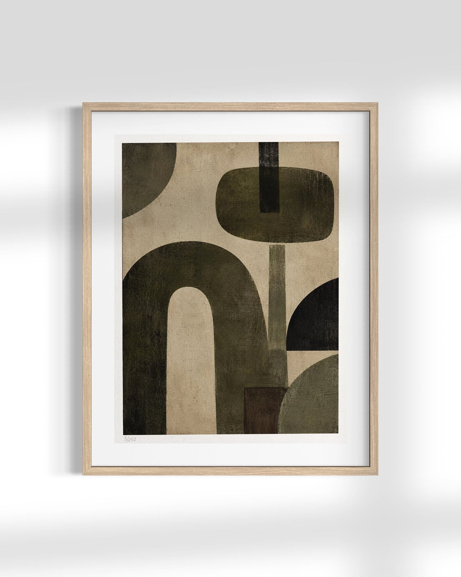 A framed abstract art print featuring organic shapes in dark olive green, black, and beige on a white mat, housed in a light natural wood frame and displayed against a light gray wall.