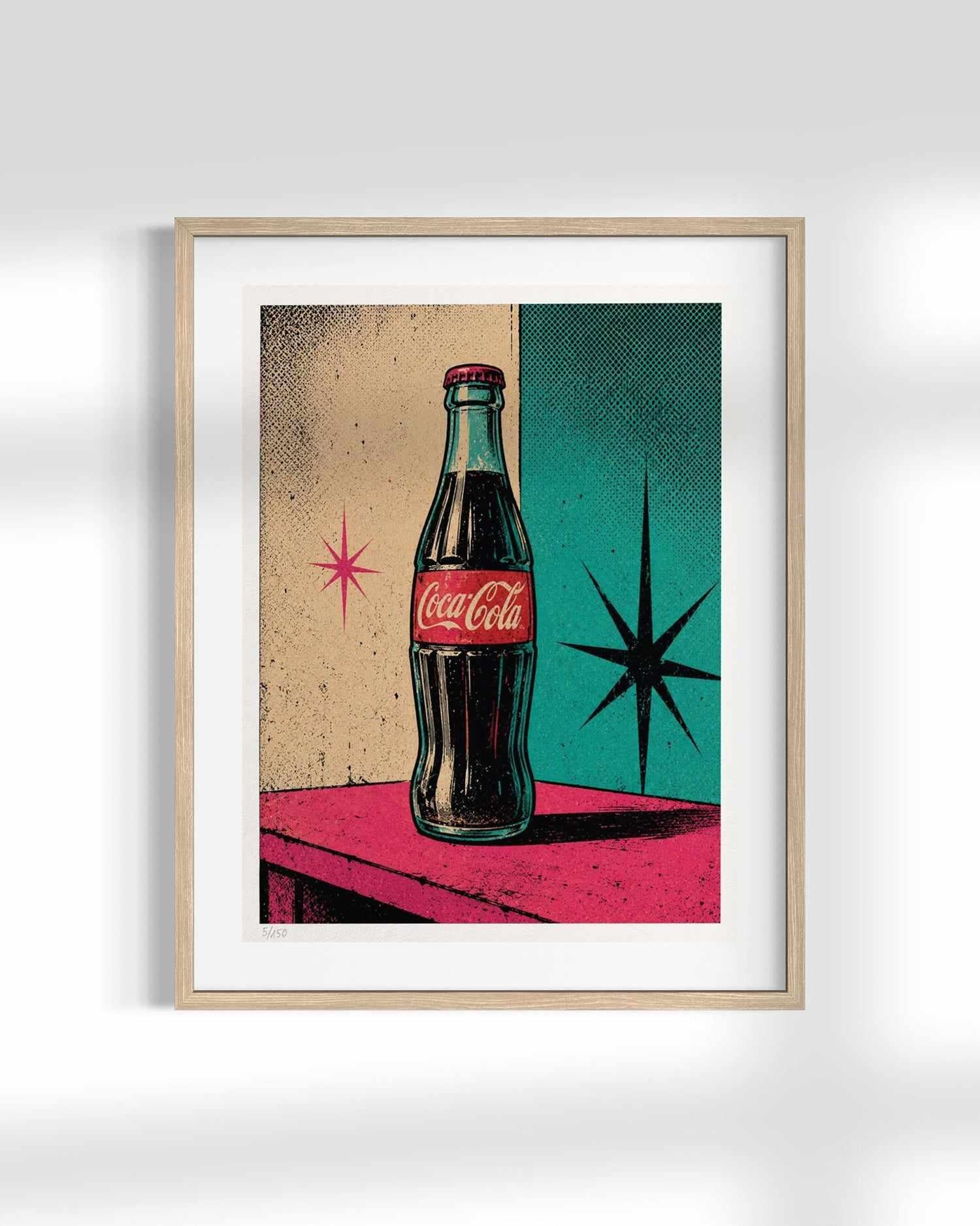A framed retro art print featuring a vintage Coca-Cola glass bottle on a pink table against a split beige and teal background with starburst shapes, shown on a light gray wall.