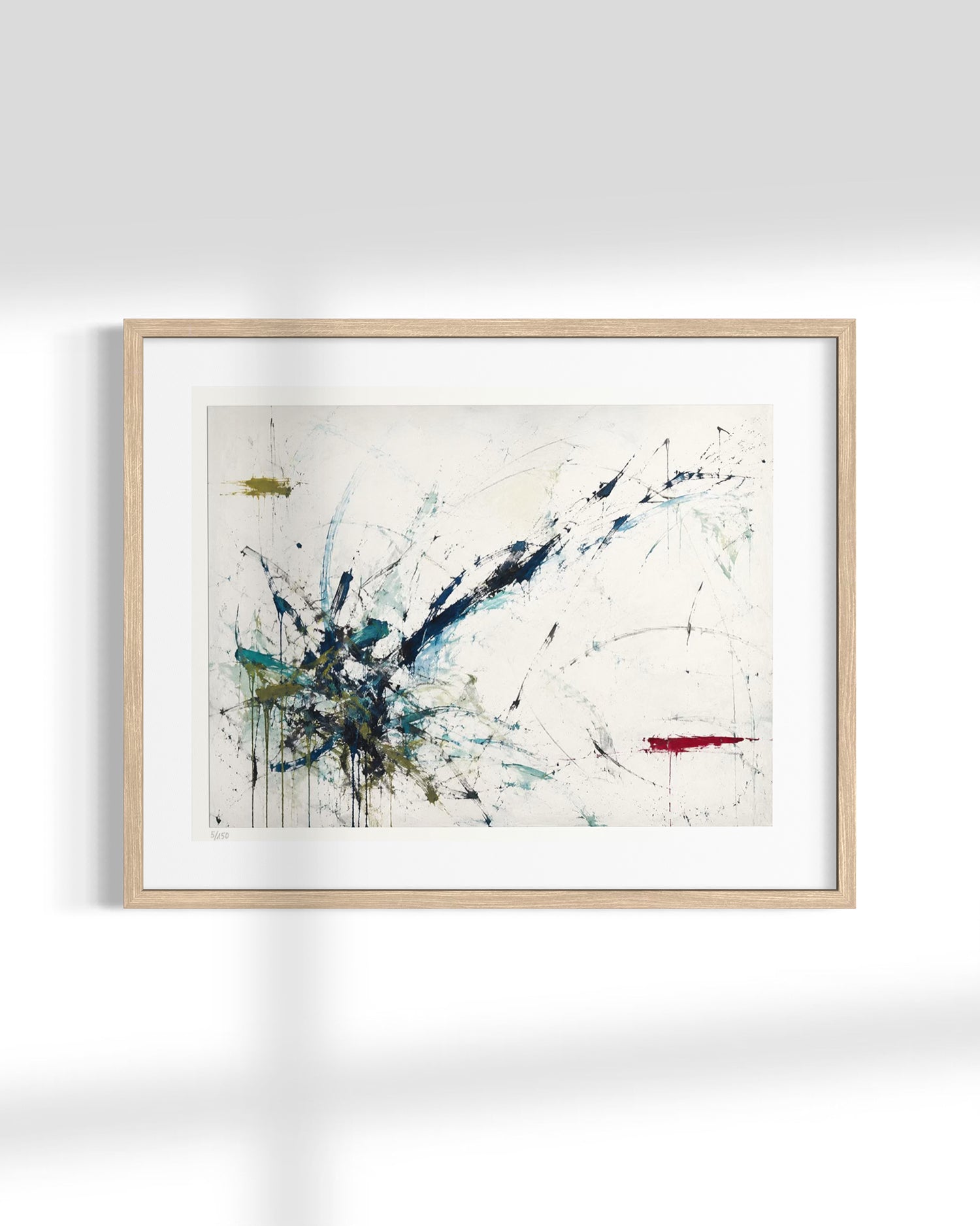 Framed abstract wall art featuring expressive blue, black, teal, and red paint splatters on an off-white background, housed in a light natural wood frame with a white mat, shown hanging on a white wall.