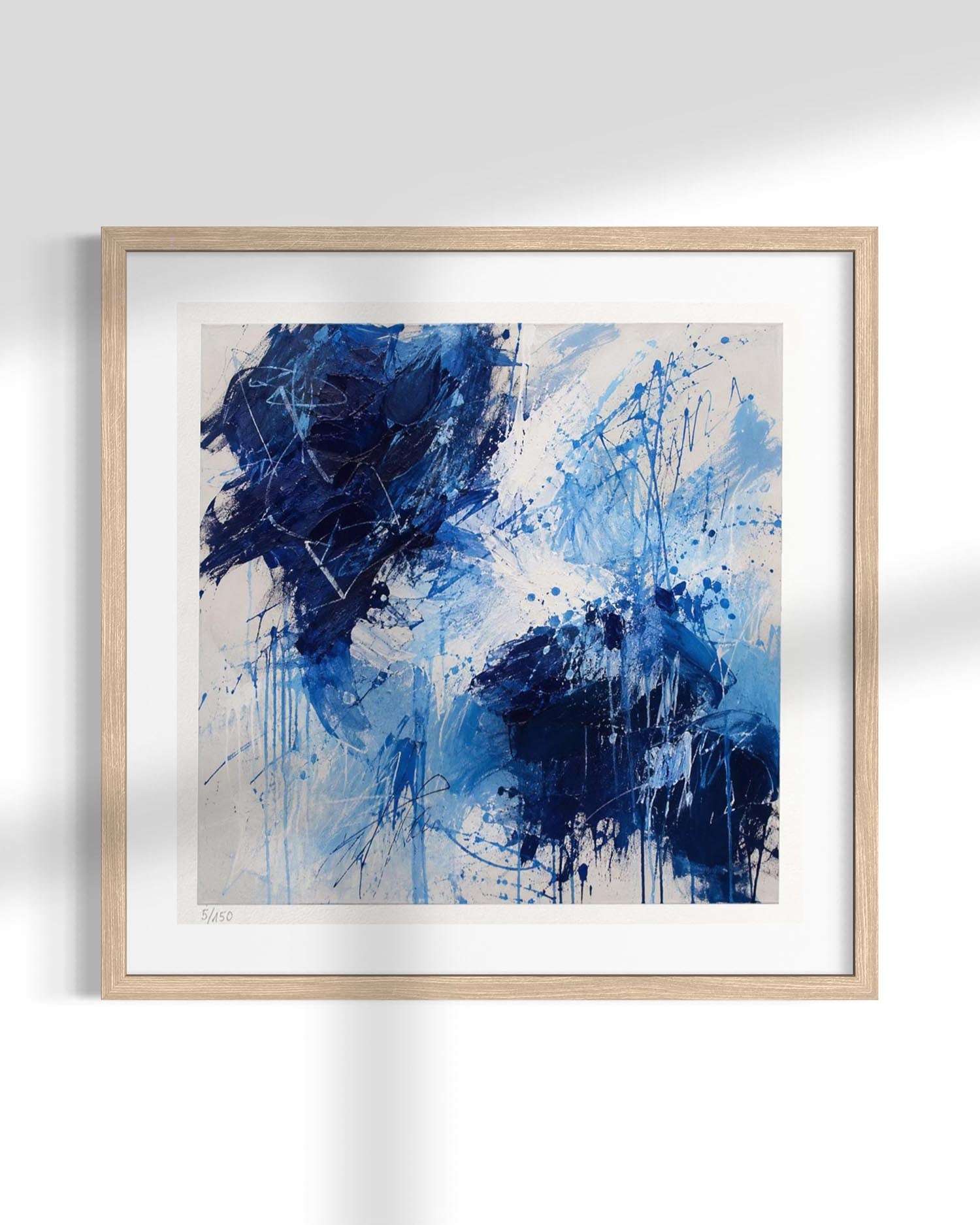 Framed abstract wall art featuring expressive blue and navy brushstrokes and splatters on a light beige background, presented in a light natural wood frame with a white mat against a white wall.
