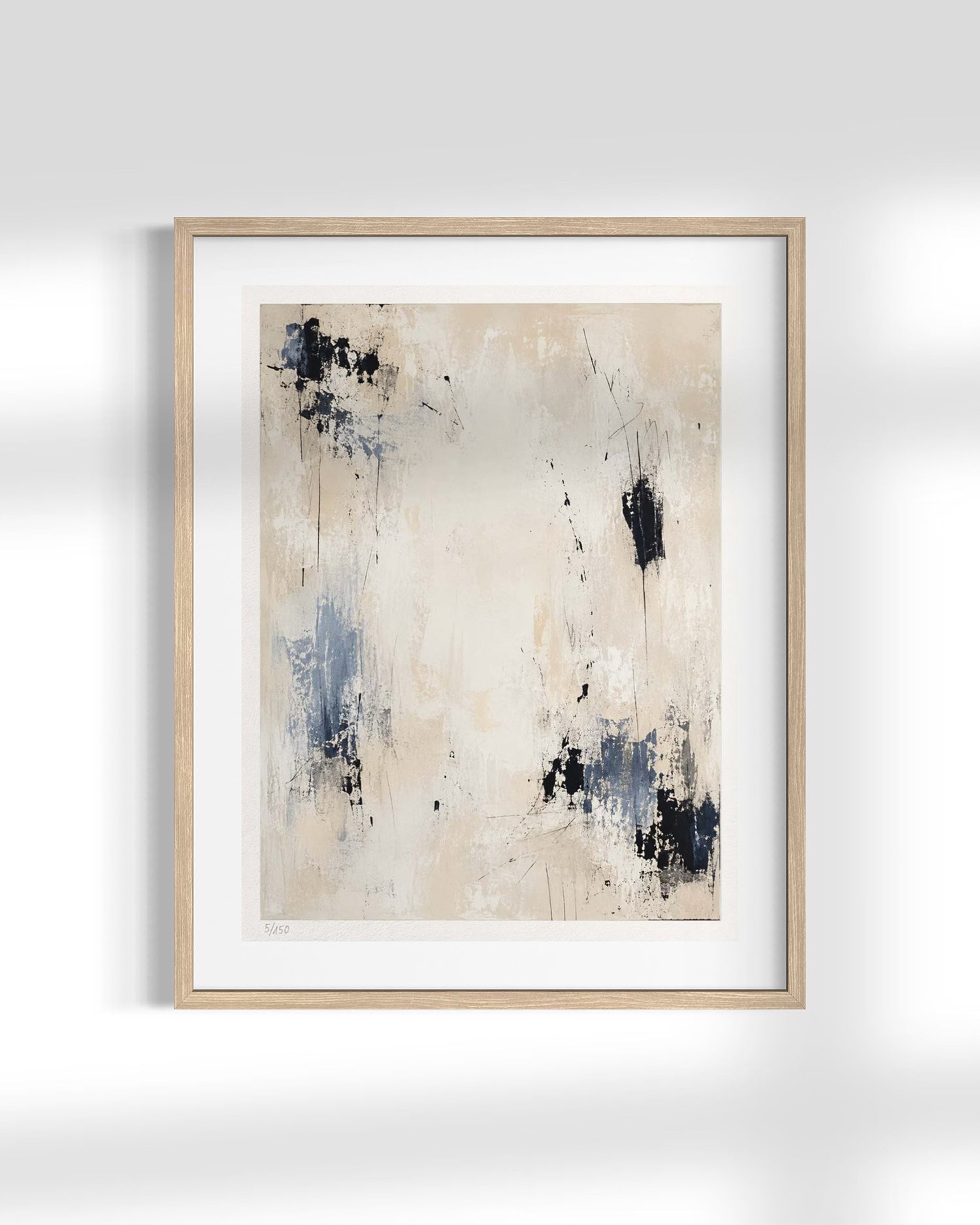 A framed abstract art print featuring a textured cream background with blue and black paint splatters, set in a light wood frame with a white mat against a white wall.