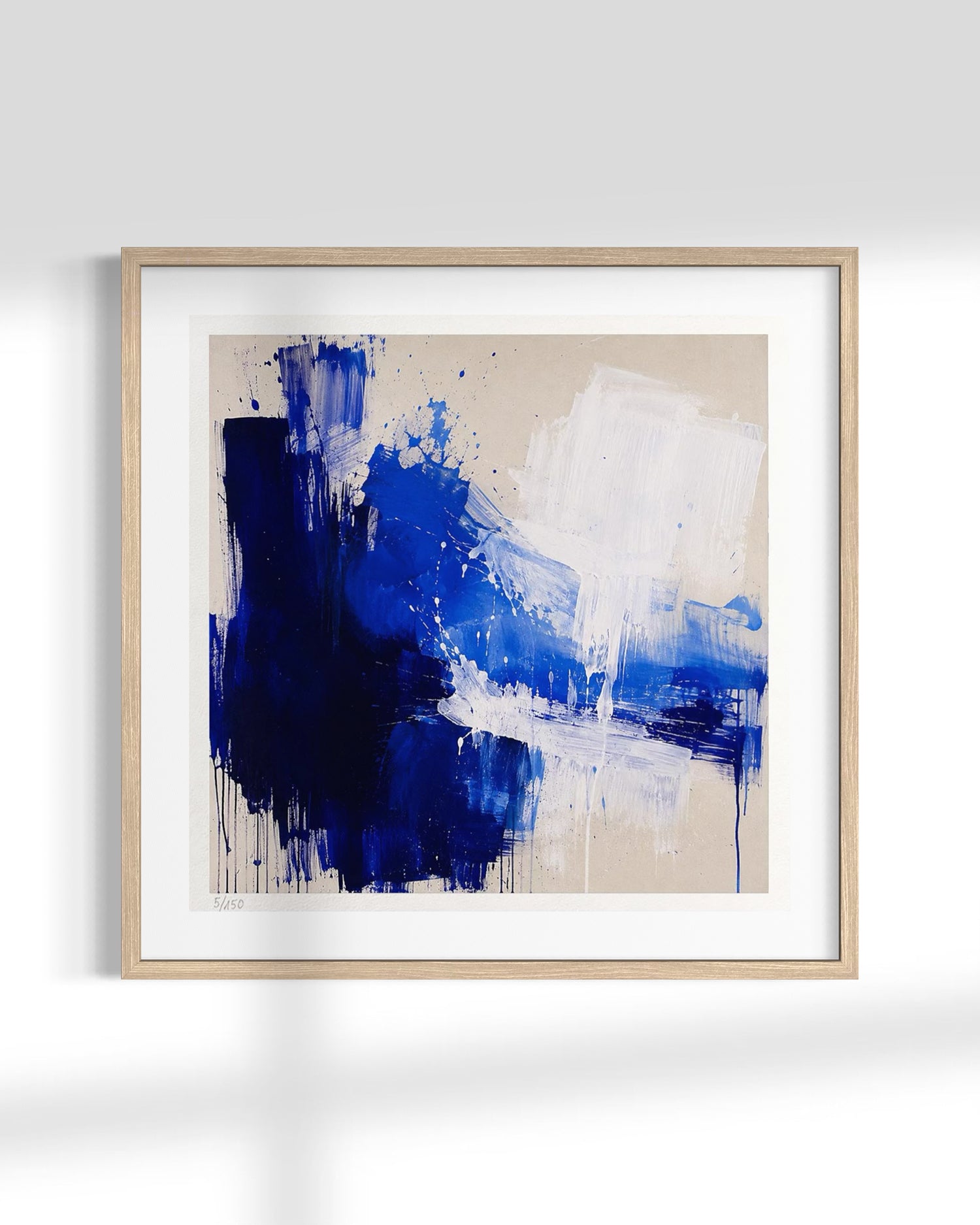 Framed abstract wall art featuring bold blue and white paint strokes on a warm beige background, presented in a light natural wood frame with a white mat against a clean white wall.