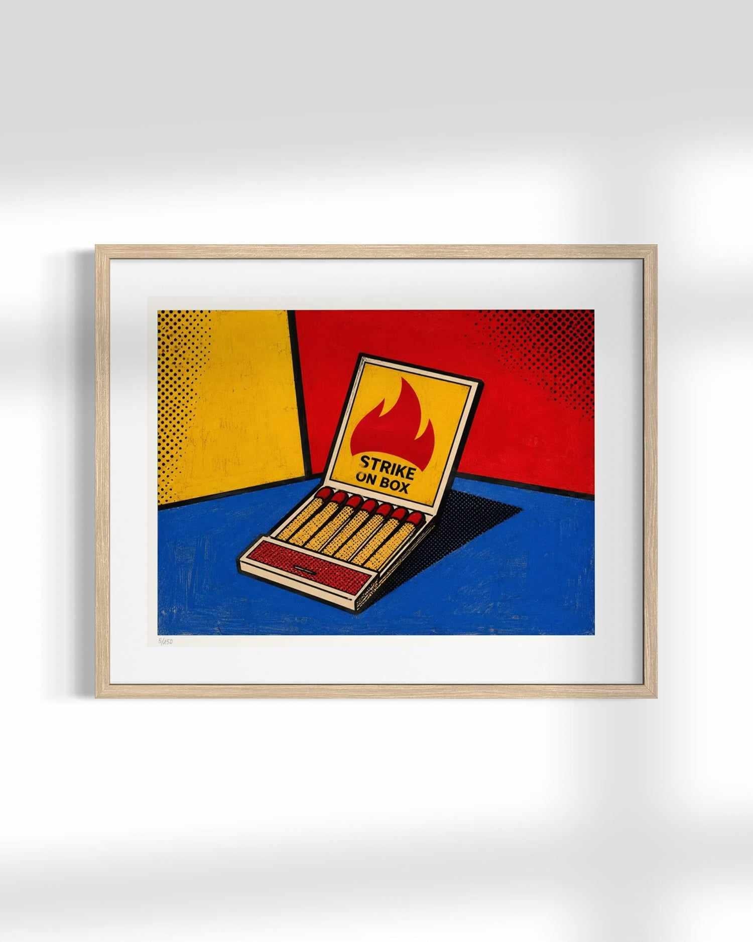 A framed pop art print depicting an open matchbox with a flame label reading 'STRIKE ON BOX', set against a bold red, yellow, and blue background. The print is housed in a light wood frame with a white mat, displayed against a neutral gray wall with soft shadows.