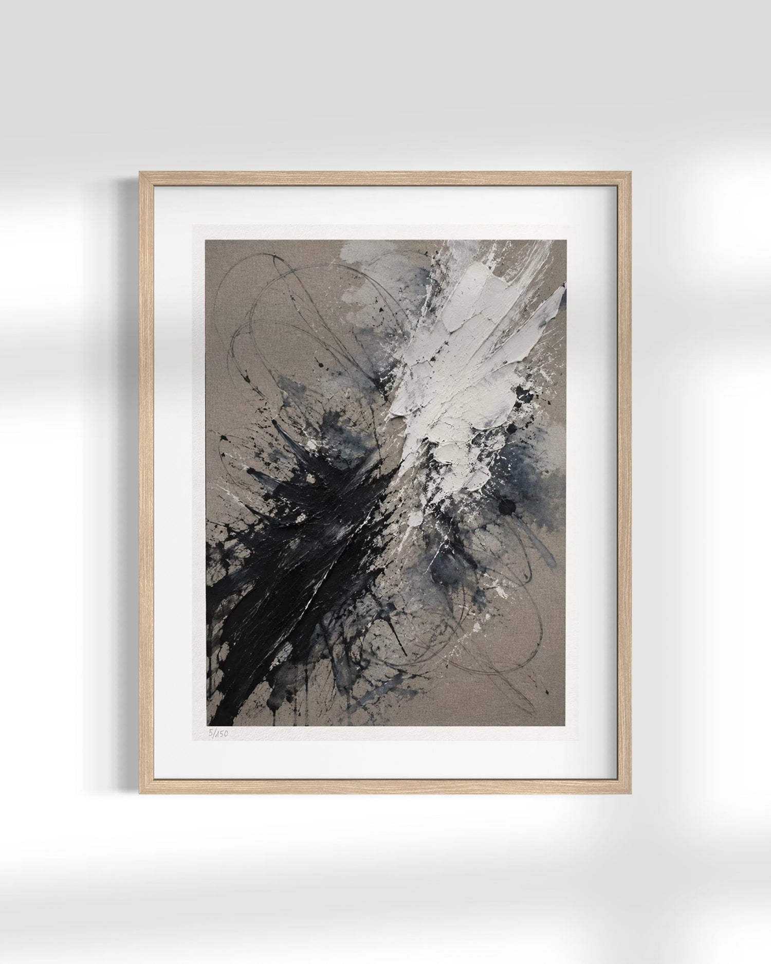 A framed abstract art print featuring black, gray, and white paint splatters on a textured taupe background, housed in a light natural wood frame with a white mat, displayed against a light gray wall.