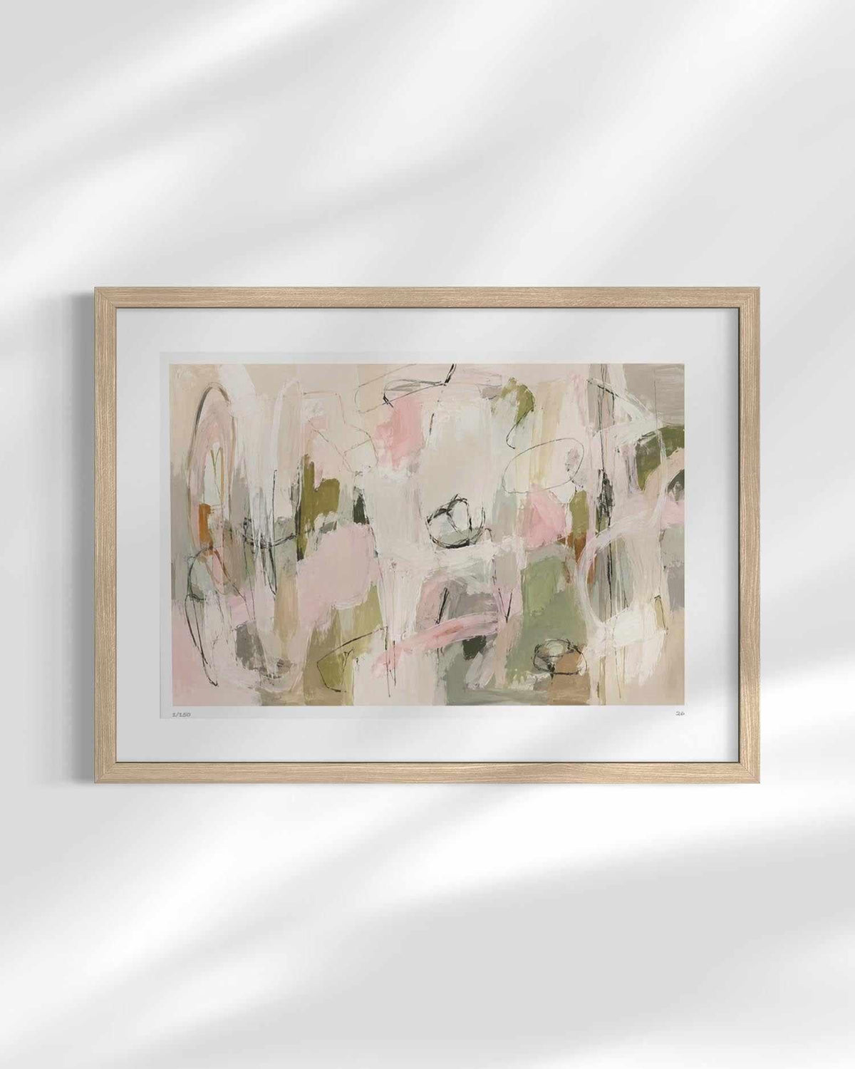 A framed abstract art print featuring soft pastel tones of pink, beige, sage green, and gray, presented in a light natural wood frame with a white mat against a white wall with soft leaf shadows.
