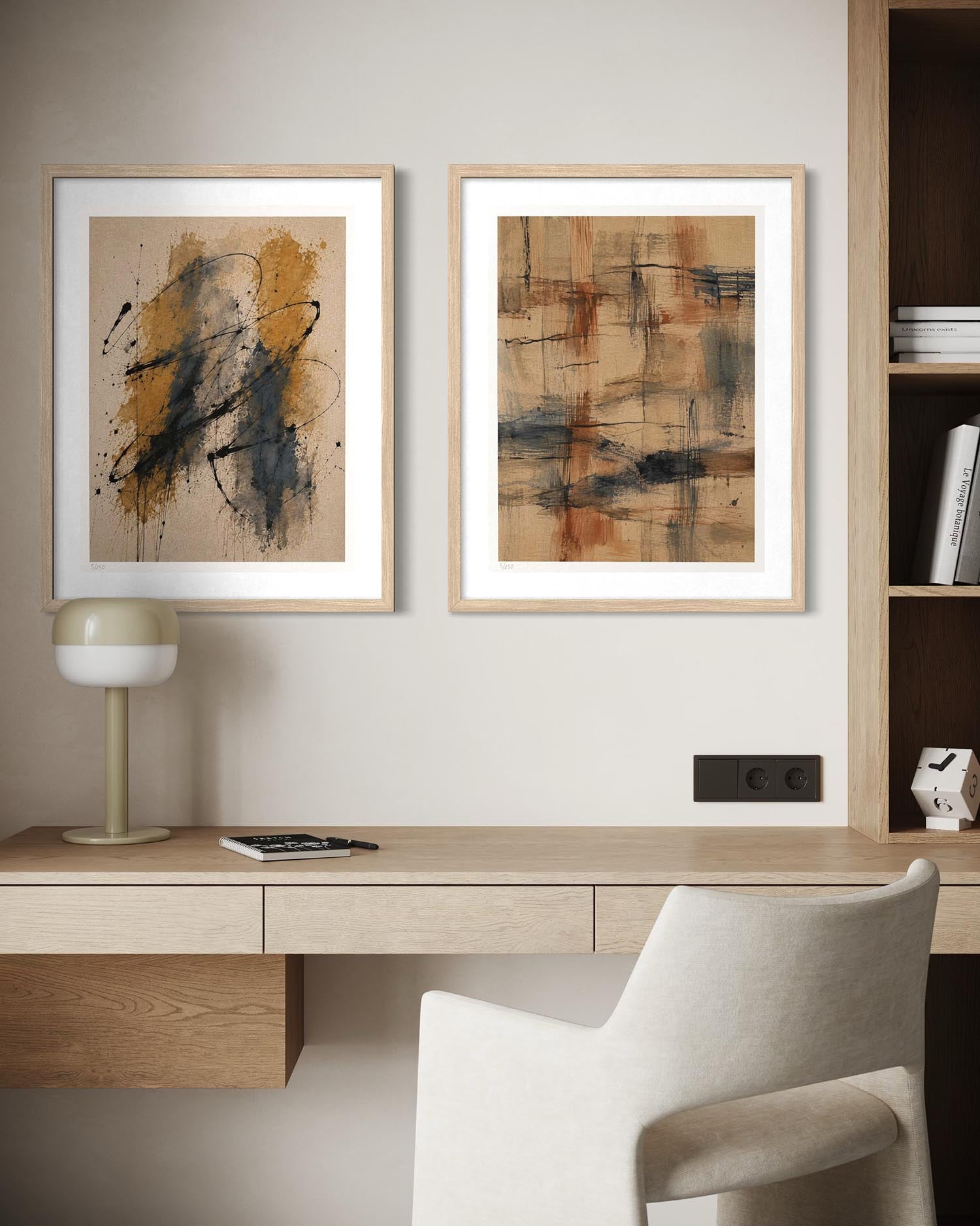 Two framed abstract art prints with light wood frames hang on a beige wall above a wooden desk with a white chair, a small table lamp, and a smartphone.
