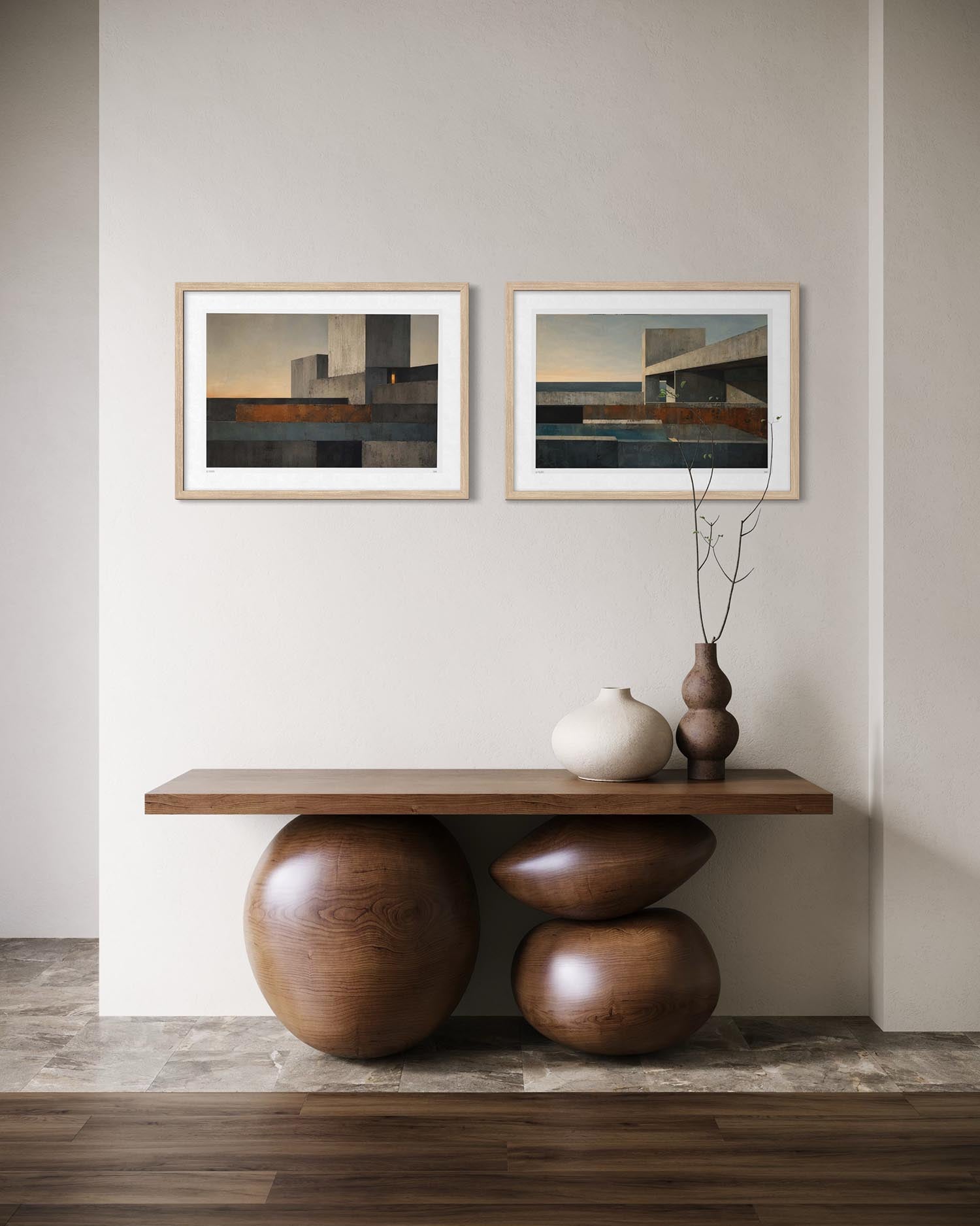 Two framed abstract landscape prints hang on a light beige wall above a wooden console table with sculptural wooden bases. The room includes a white ceramic vase and a dark brown vase with dried branches on the table.
