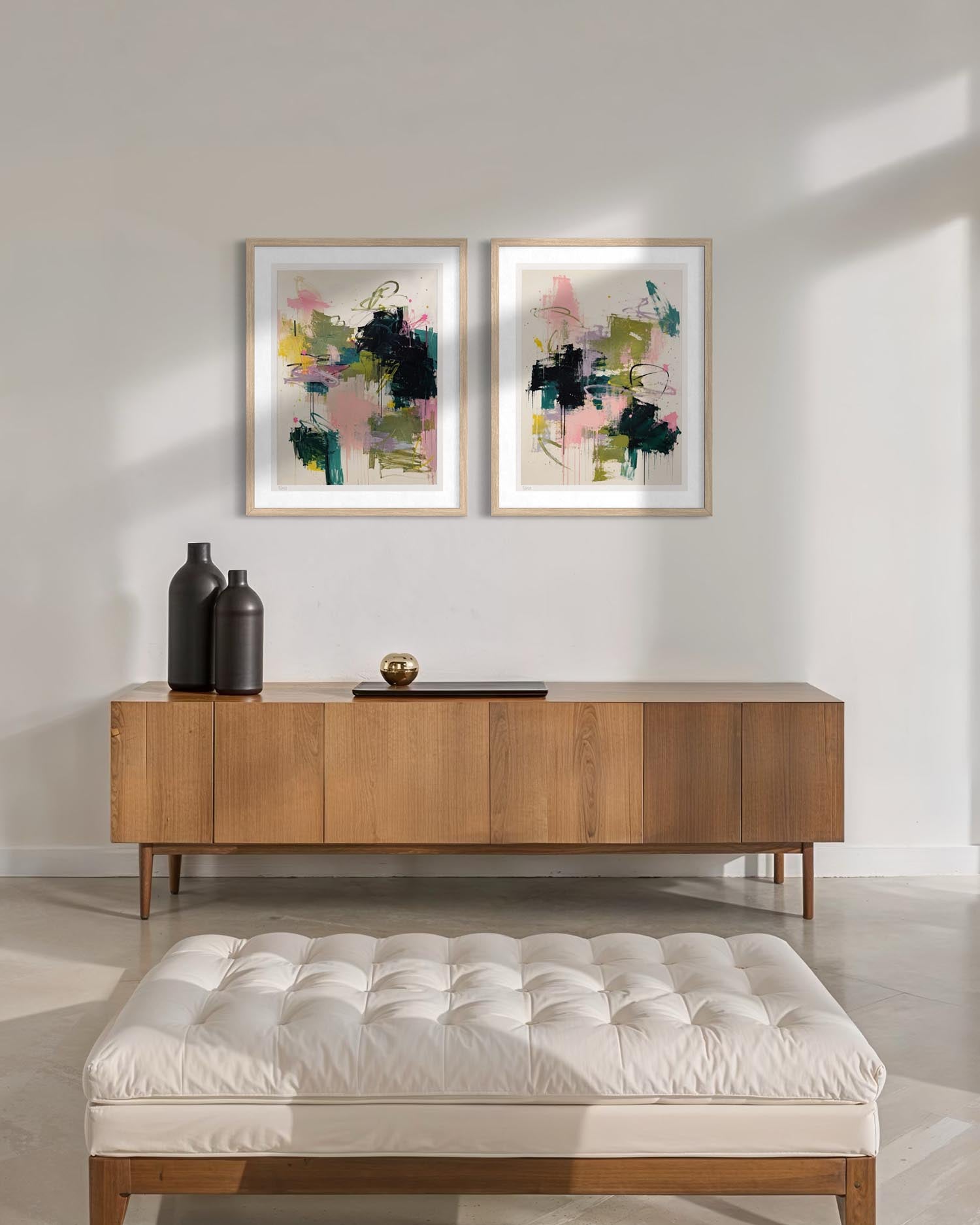 A styled interior scene with two framed abstract wall art prints hanging above a mid-century modern wooden sideboard. The room has a white wall, light floor, a white tufted bench in the foreground, and two dark bottles on the sideboard.