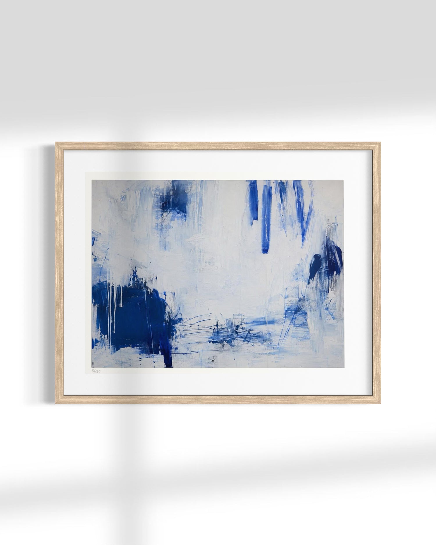 A framed abstract art print featuring blue and white brushstrokes, presented in a light natural wood frame with a white mat against a white wall.