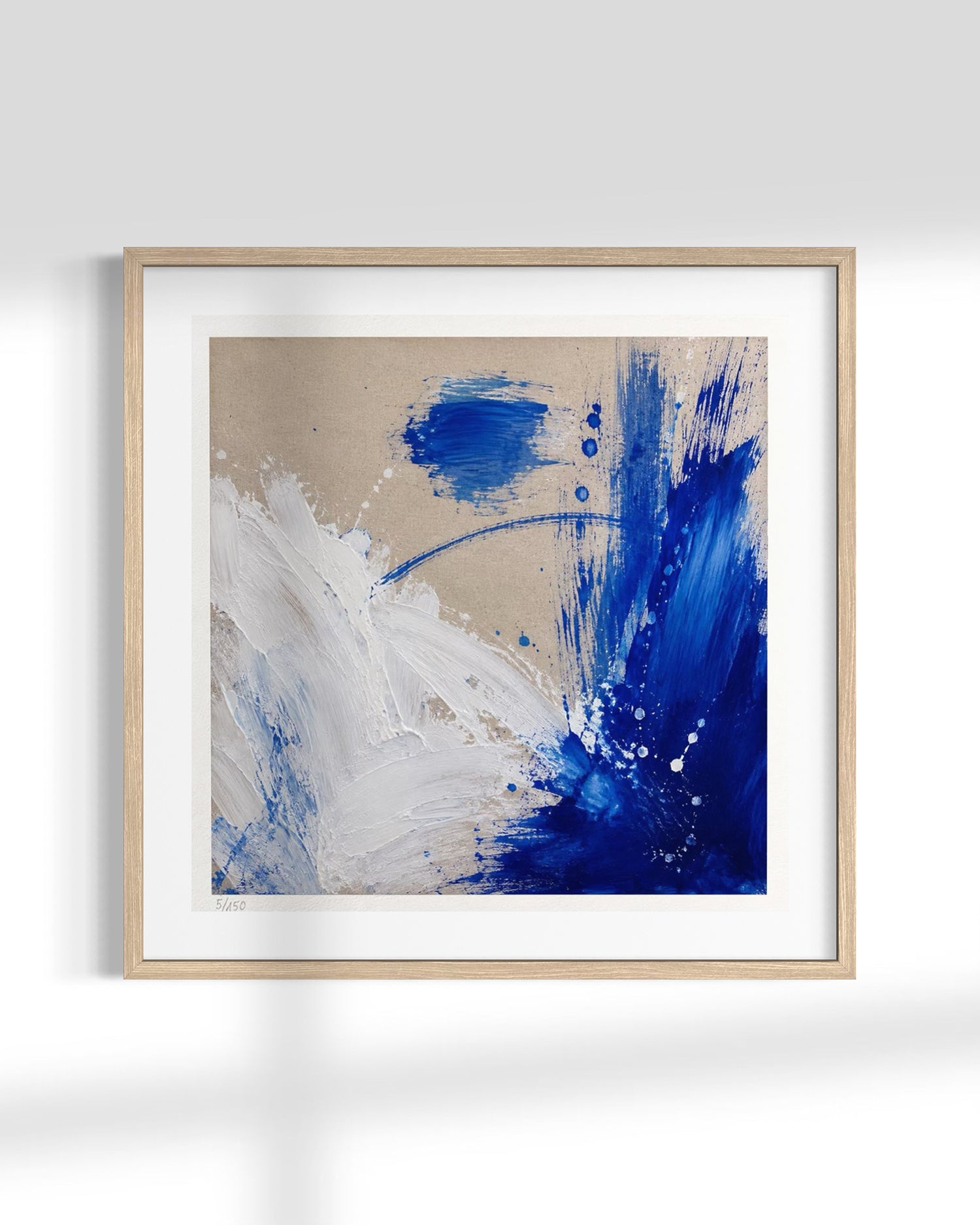 Framed abstract wall art featuring bold blue and white paint strokes on a warm beige background, presented in a light natural wood frame with a white mat against a clean white wall.