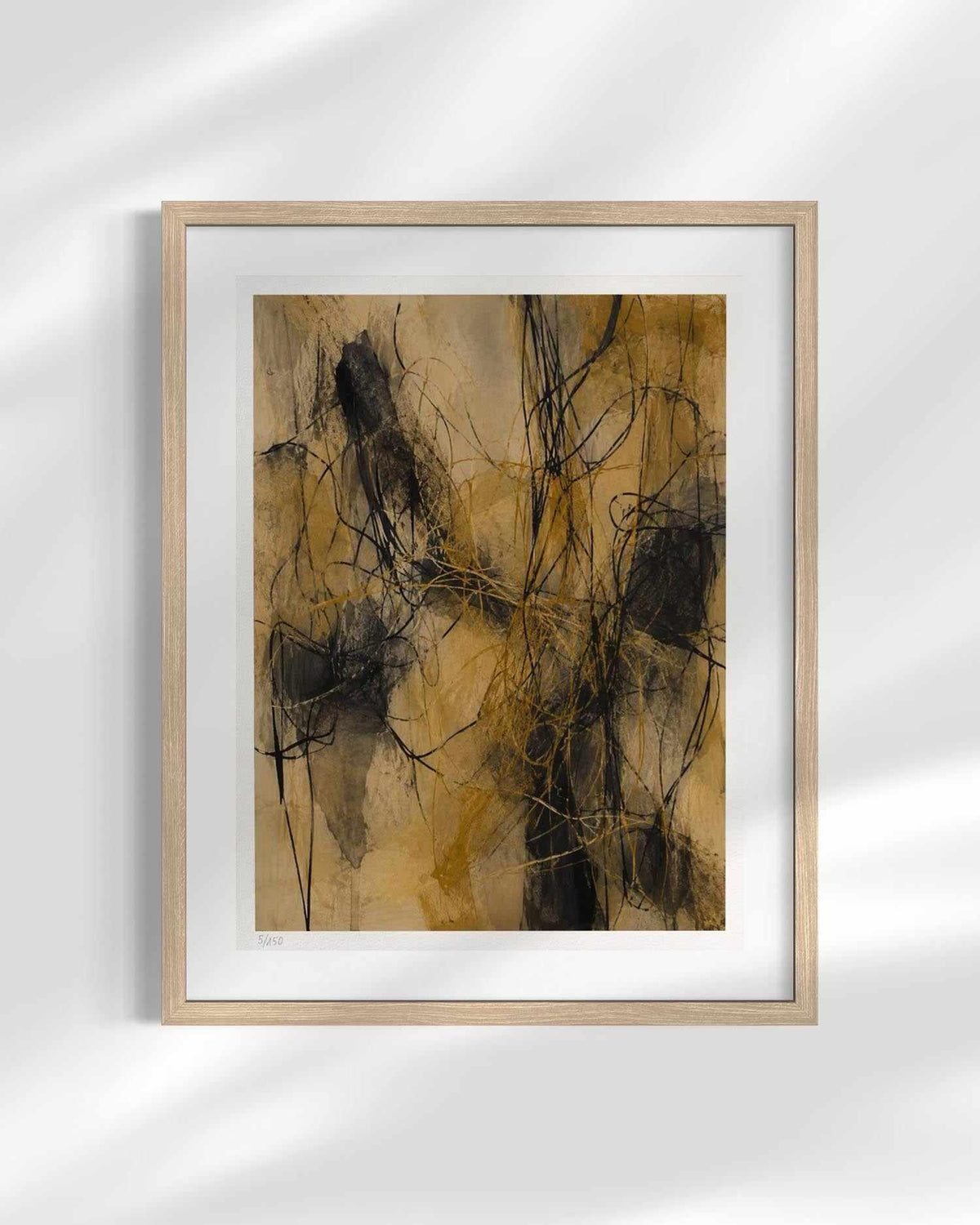 Framed abstract wall art featuring expressive black and dark brown brushstrokes over a warm tan and beige background, presented in a light natural wood frame with a white mat against a softly lit gray wall.