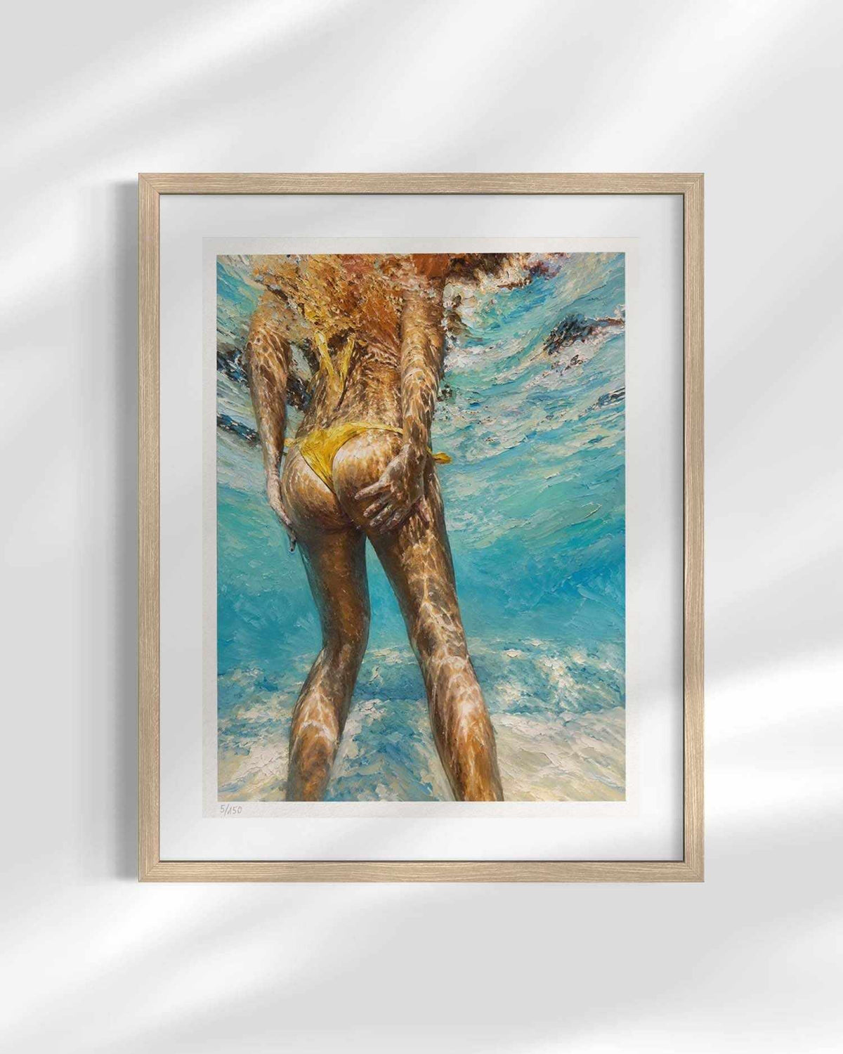 A framed art print featuring an underwater painting of a figure in a yellow bikini, set in a light wood frame with a white mat against a white wall with soft shadow overlays.