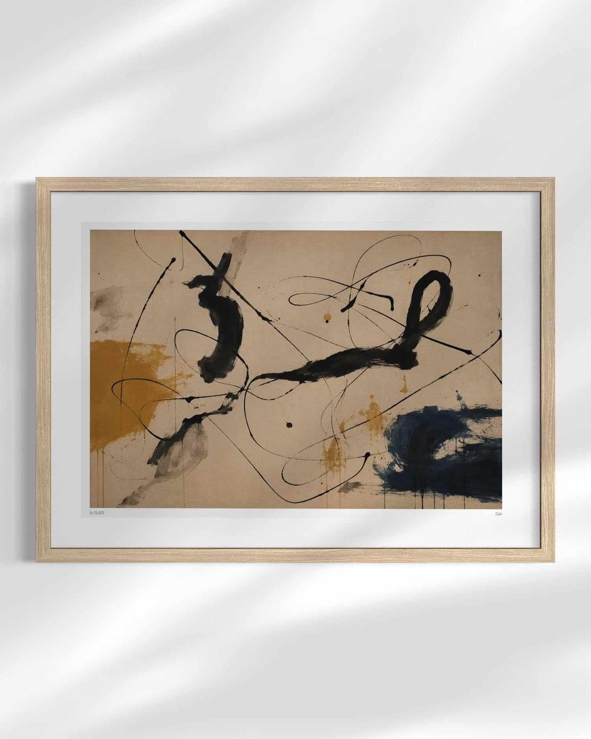 A framed abstract art print featuring black and navy brushstrokes and splatters on a beige background, enclosed in a light wood frame with a white mat, shown against a white wall with soft leaf shadows.