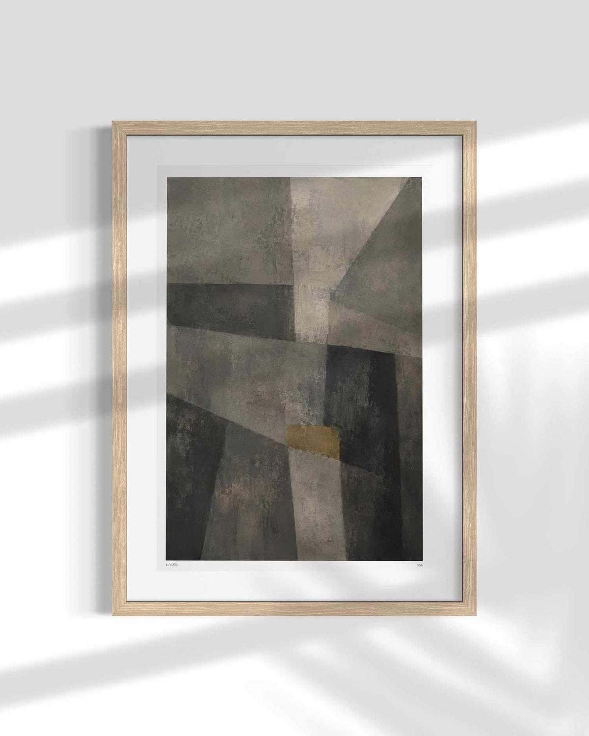 A framed abstract art print featuring geometric shapes in shades of gray, brown, and black with a small gold accent, displayed on a white wall with soft window shadow patterns.