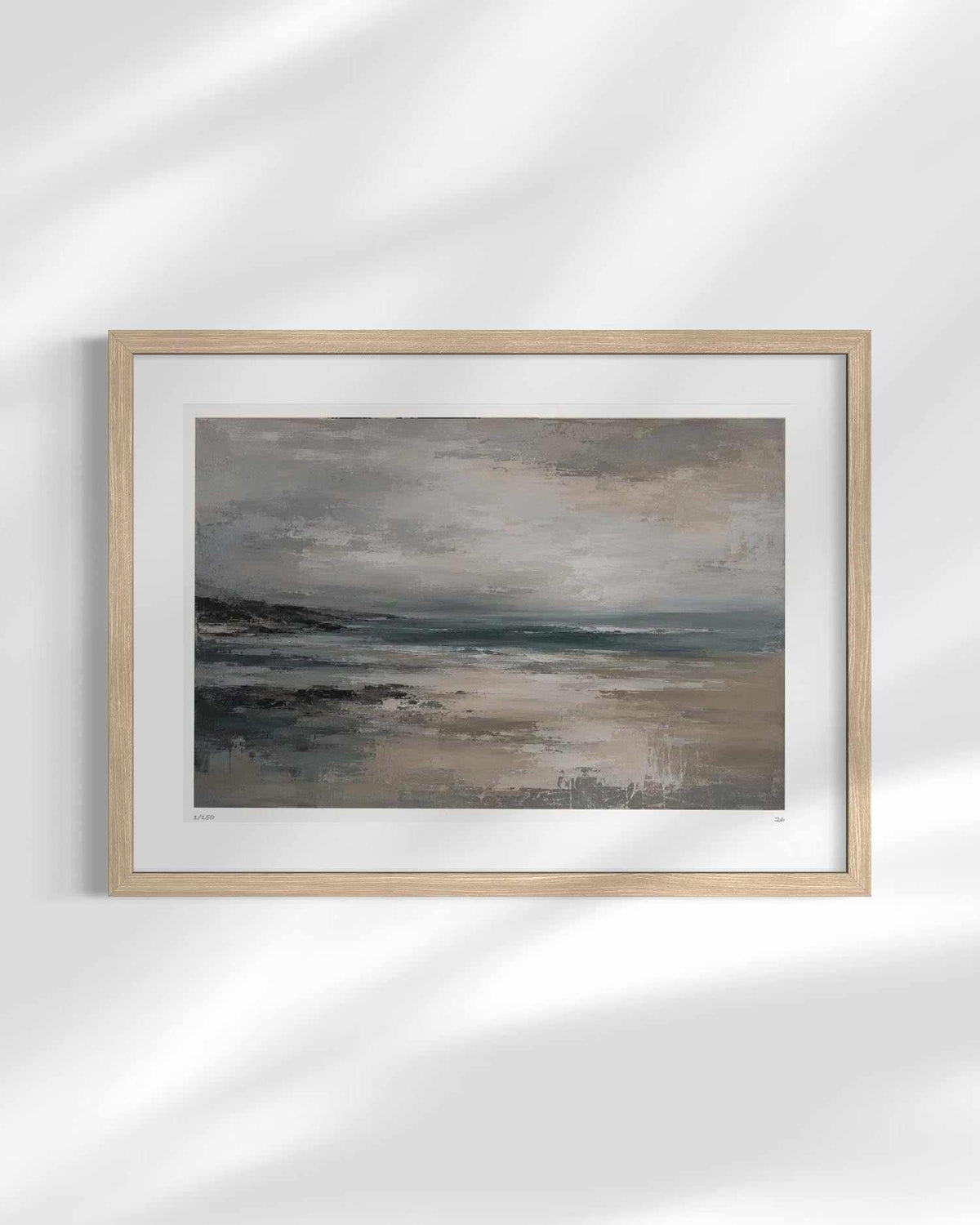 A framed landscape art print depicting a misty beach and ocean scene, mounted on a white wall with soft, diagonal shadow overlays.