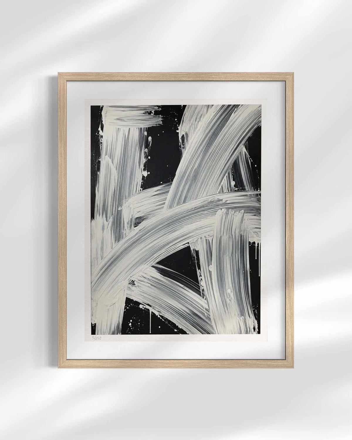 Framed abstract wall art featuring bold white and gray brushstrokes on a black background, presented in a light natural wood frame with a white mat against a softly lit gray wall.