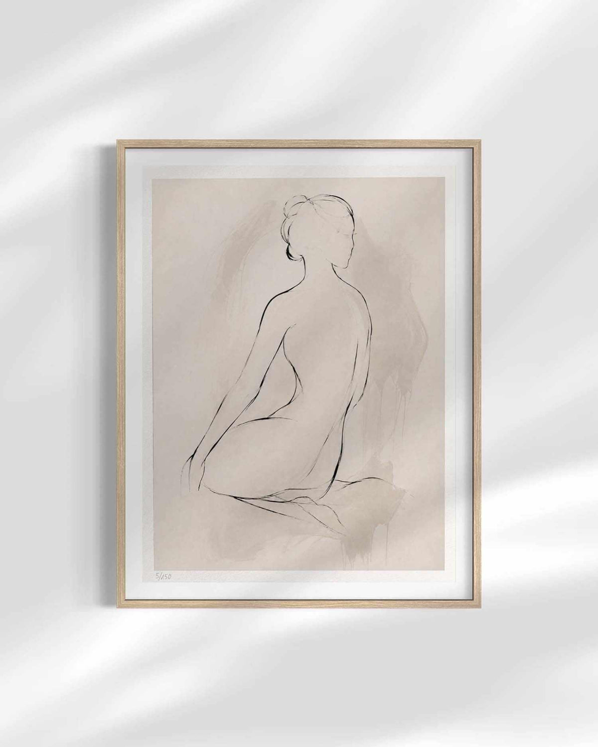 A minimalist framed art print featuring a black line drawing of a seated nude figure on a warm beige background, enclosed in a light wood frame with a white mat, displayed against a white wall with soft shadow overlays.