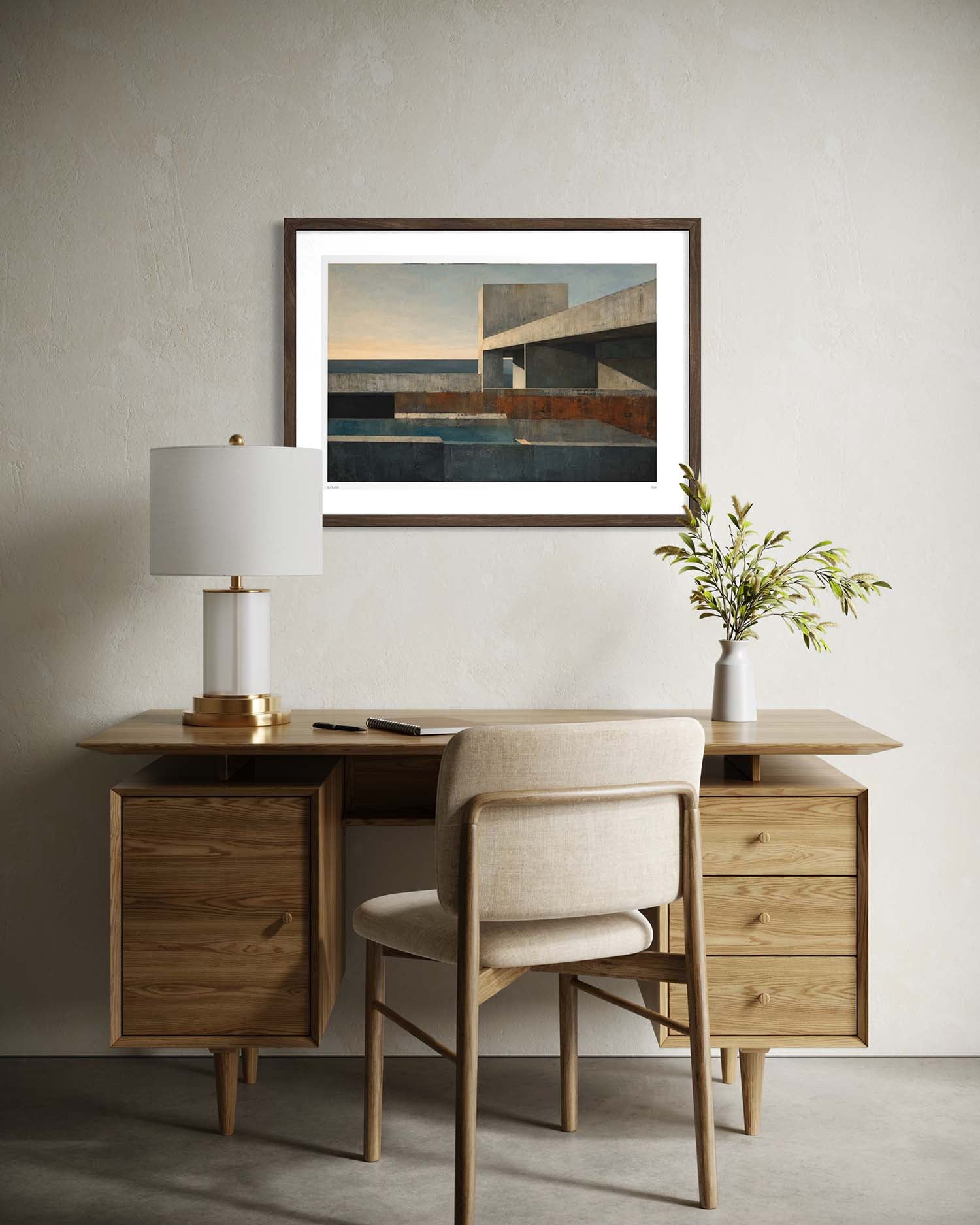A framed abstract landscape art print hangs on a textured light gray wall above a mid-century modern wooden desk and chair. The print features geometric structures in shades of blue, brown, and beige under a warm sunset sky.