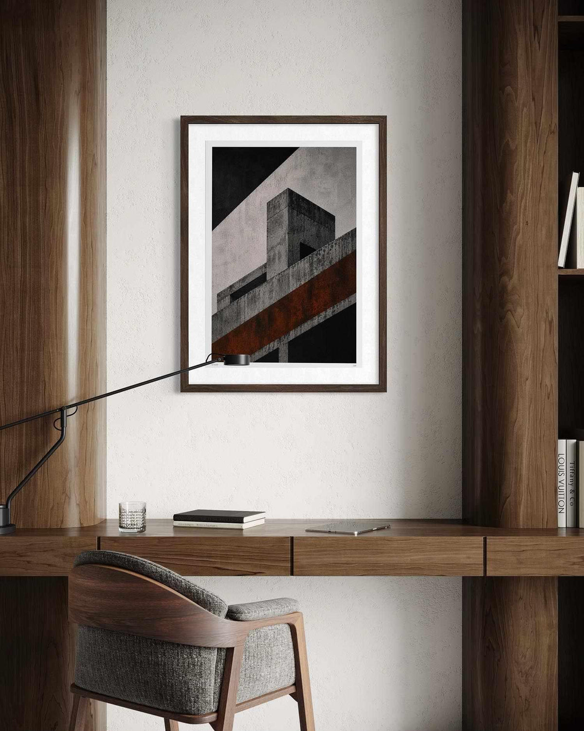 A framed abstract architectural art print with black, white, gray, and rust-orange tones, displayed on a light wall above a wooden desk with a chair and lamp.