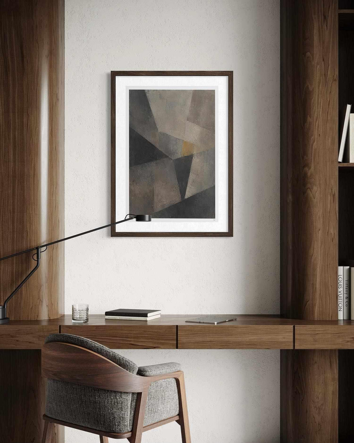 A framed abstract geometric art print with shades of gray, brown, and beige, displayed on a light wall above a wooden desk with a chair and a desk lamp.