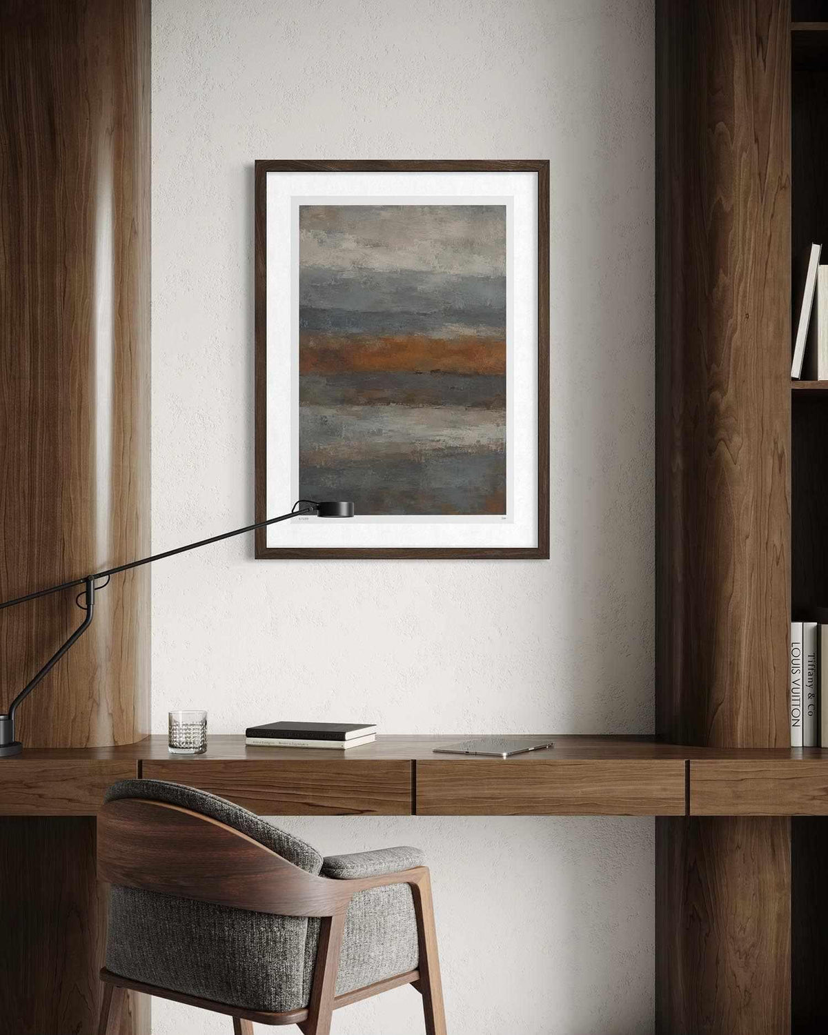 A framed abstract landscape art print hangs on a light beige wall above a wooden desk with a chair and a modern desk lamp. The artwork features horizontal bands of gray, blue, and a warm brown-orange.