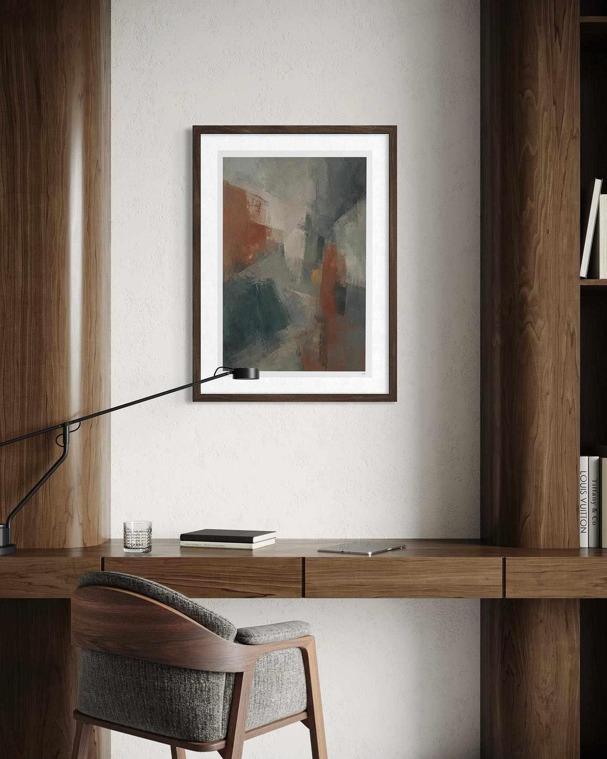 A framed abstract art print with a dark brown frame and white mat, hanging on a light gray wall above a wooden desk with a chair and lamp.