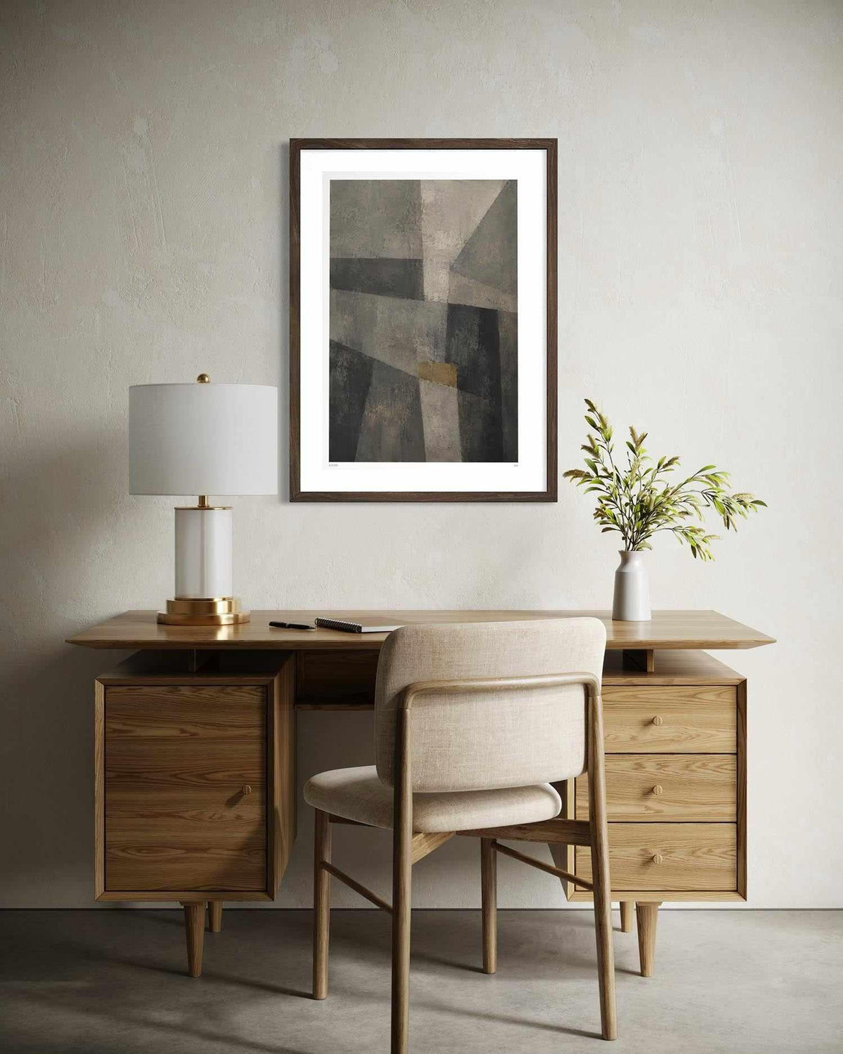 A framed abstract art print with a dark wood frame and white mat, hanging on a textured light gray wall above a mid-century modern wooden desk and chair.