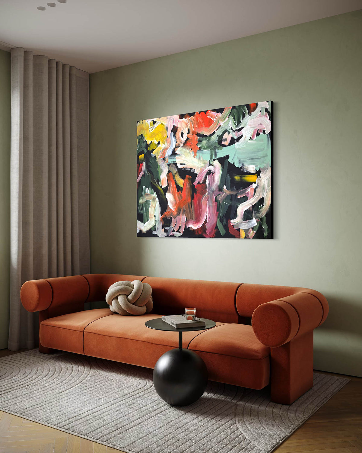 An abstract canvas painting with vibrant multicolor brushstrokes hangs on a sage green wall above a modern rust-orange velvet sofa in a styled living room.