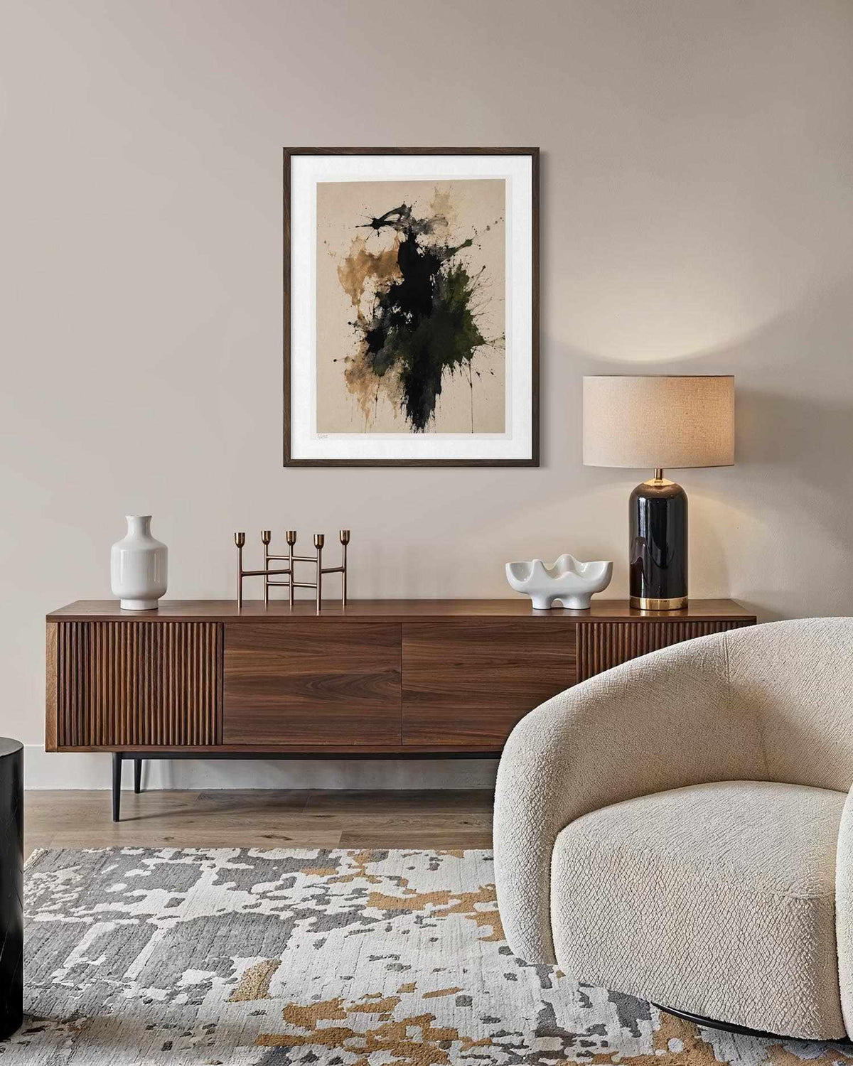 A framed abstract art print hangs on a beige wall above a mid-century modern wooden sideboard in a styled living room. The artwork features a dark green and black brushstroke design on a textured beige background.