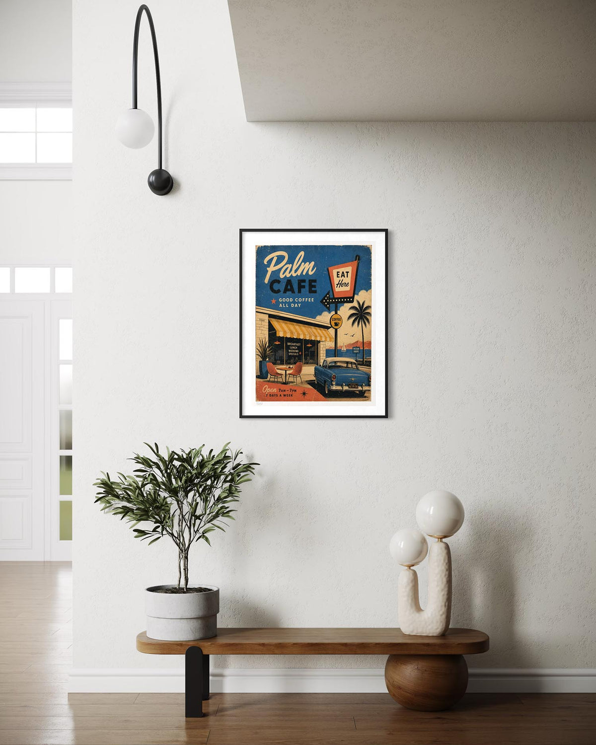 A framed vintage-style wall art print reading 'Palm Cafe' hangs on a white wall above a wooden bench with a potted plant and a sculptural decor object in a modern interior setting.