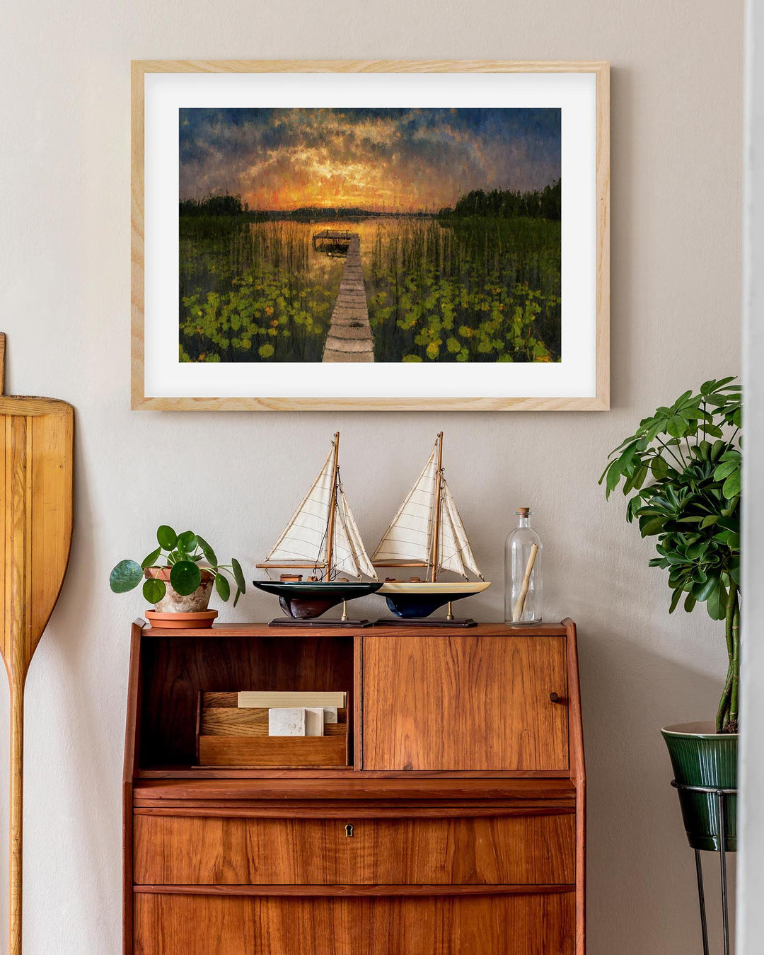 Framed artwork of a sunset over water with a wooden cabinet below displaying decorative items.