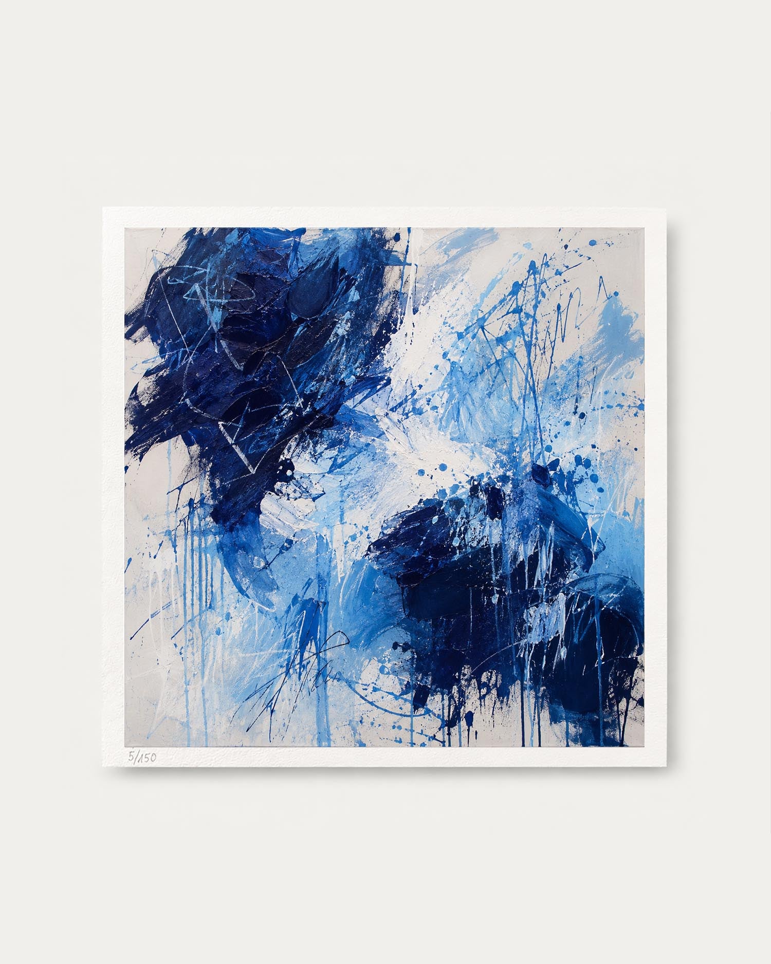An abstract square canvas print featuring expressive blue and navy paint splatters and drips on a light off-white background, framed by a clean white border against a neutral light gray wall.