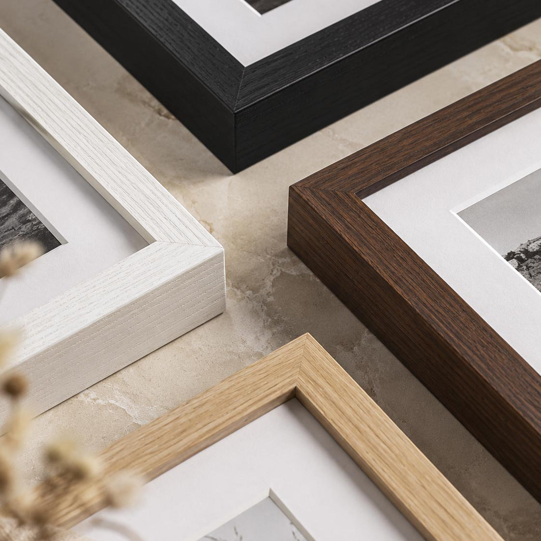 Four picture frames in different wood tones and finishes arranged on a light stone surface: black, dark brown, light natural wood, and white. White matting is visible inside the frames, with a small dried flower in the lower left corner.