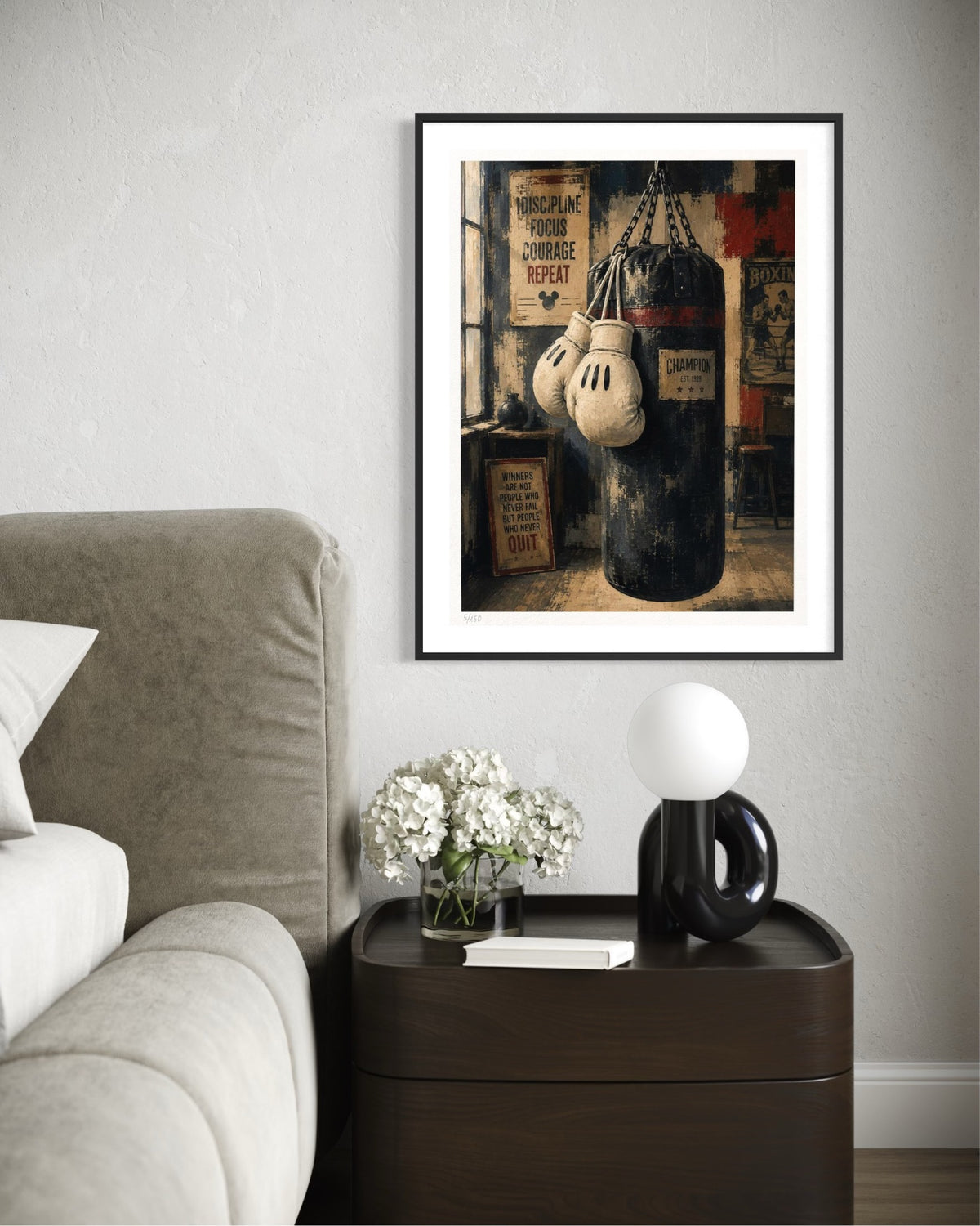 A framed art print depicting a vintage boxing scene with a punching bag and boxing gloves, styled on a light gray wall above a dark wood nightstand with a lamp and flowers.