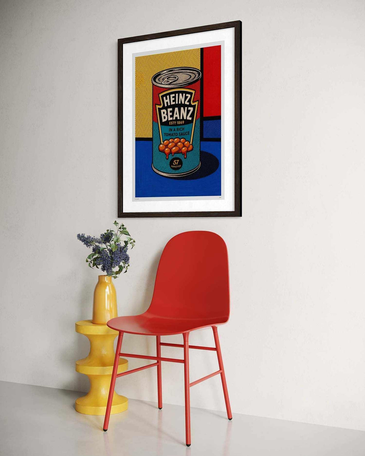 A framed pop art print of a Heinz Bean can hanging on a white wall above a red chair and a yellow side table with flowers.