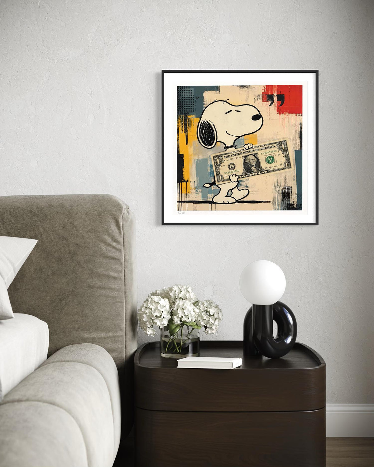 A framed wall art print featuring Snoopy holding a dollar bill, displayed in a bedroom setting above a dark wood nightstand with a lamp and flowers.