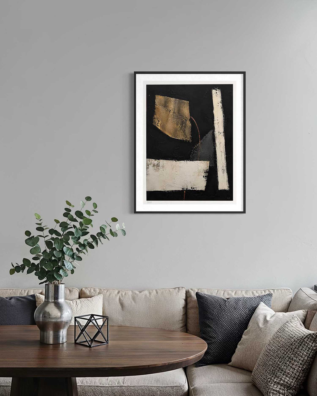 A framed abstract art print with black, gold, and cream brushstrokes hangs on a light gray wall above a beige sofa with pillows and a wooden coffee table with a vase of greenery.