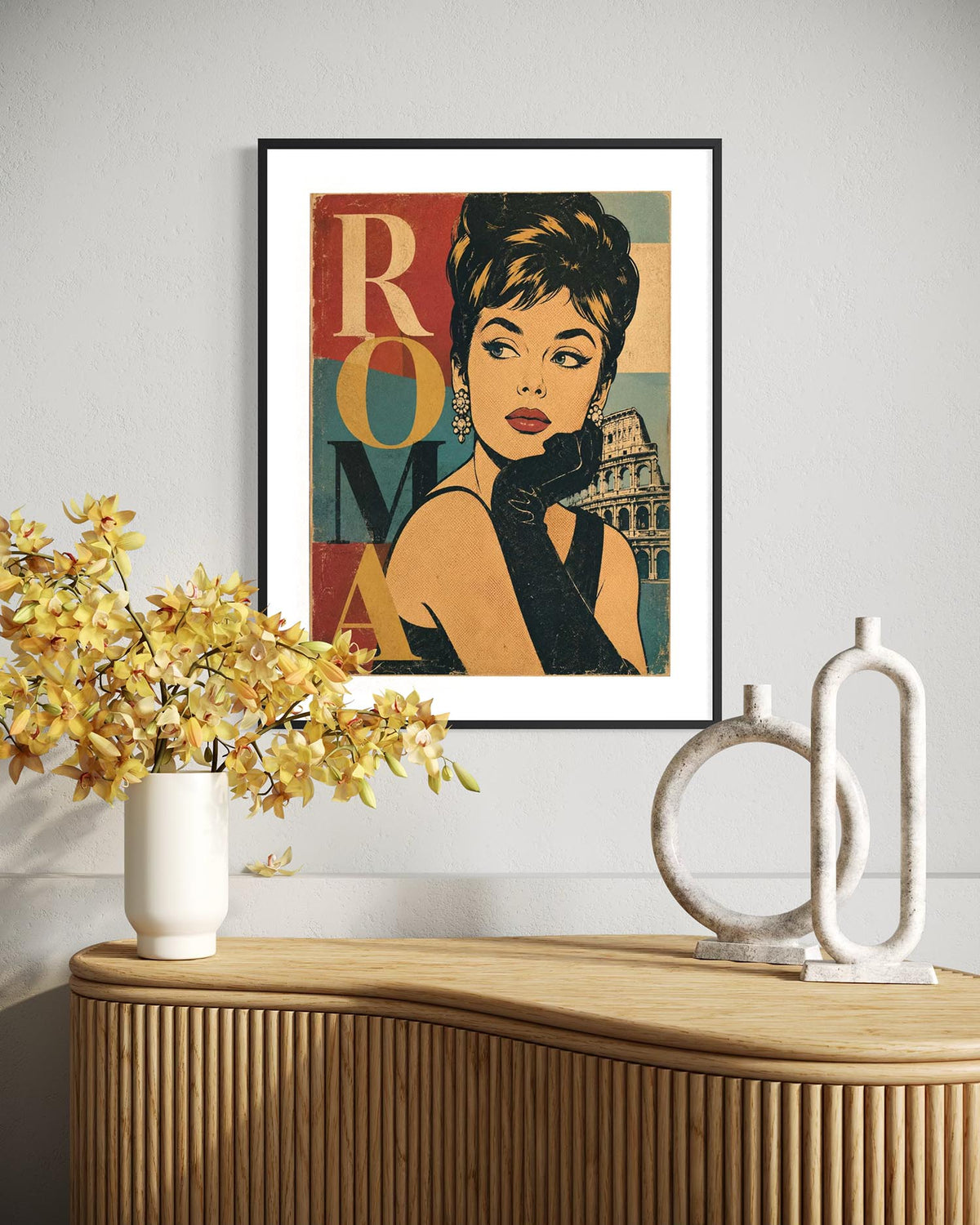 A framed pop-art poster featuring a stylized portrait of Audrey Hepburn with the word 'ROMA' and the Colosseum, displayed on a light gray wall above a wooden console table with a vase of yellow flowers and white decorative objects.