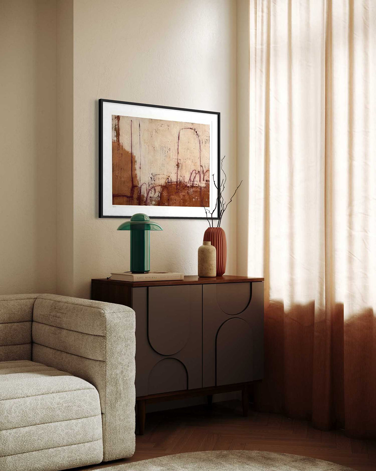 A framed abstract art print with earthy brown and beige tones hangs on a beige wall above a dark wood credenza in a styled living room corner with a sofa and sheer curtains.