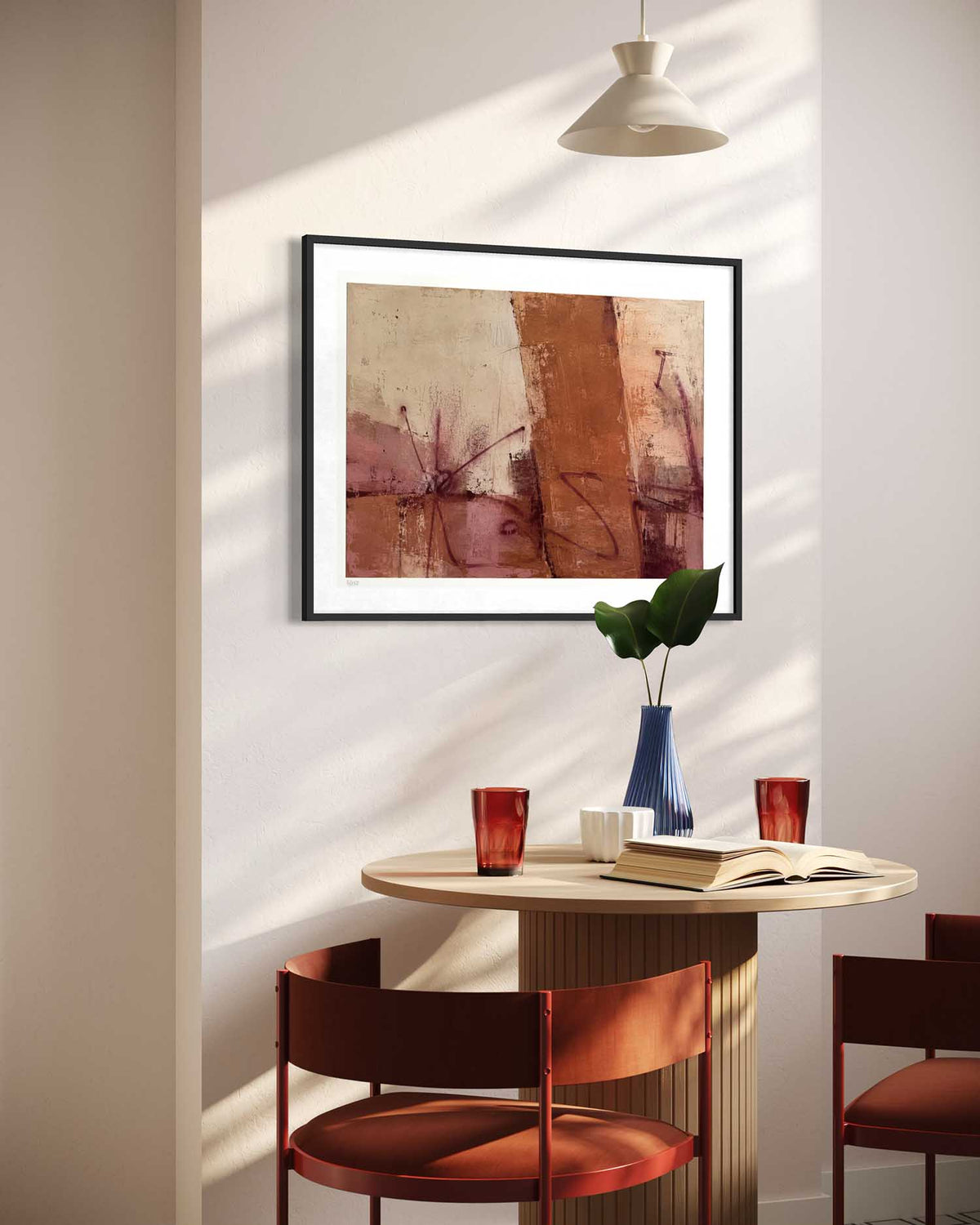 A framed abstract art print with warm terracotta, brown, and beige tones hanging on a wall in a modern dining room setting with a wooden table, chairs, and a hanging lamp.