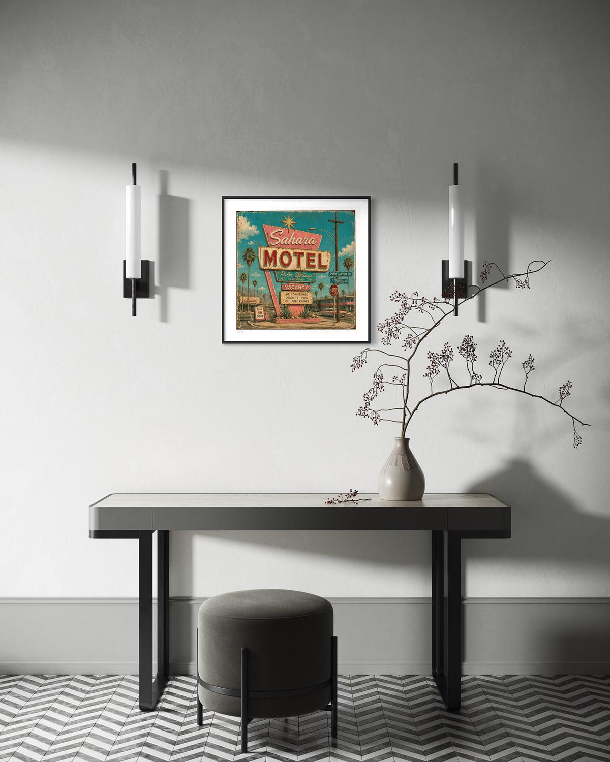 A framed vintage-style 'California Motel' wall art print hangs above a long dark console table in a modern room. The scene includes two black wall sconces, a gray upholstered stool, a ceramic vase with dried branches, and a gray-and-white chevron floor.
