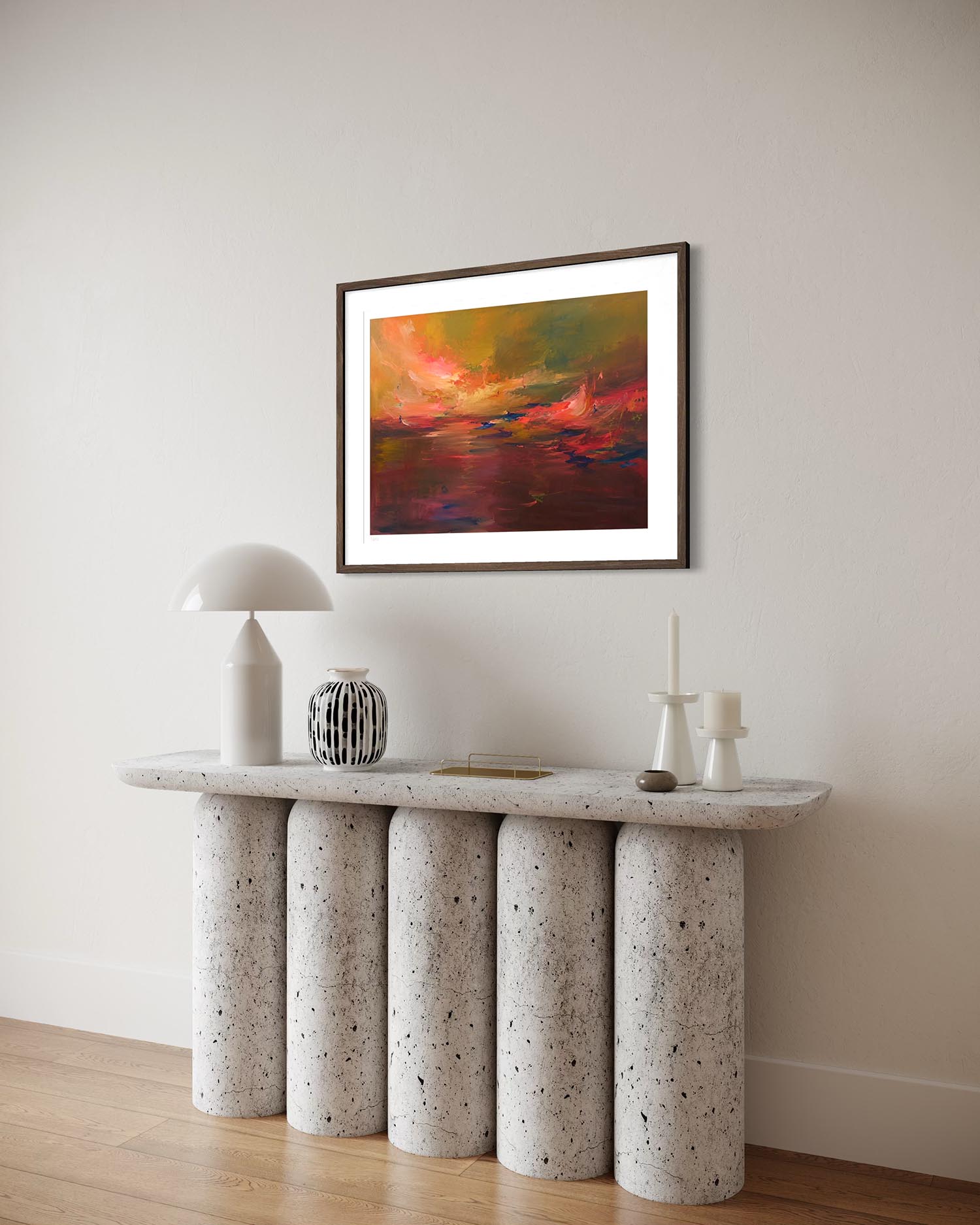 A framed abstract painting with vibrant red, orange, and yellow tones hangs on a light beige wall above a modern console table with cylindrical stone legs and a white lamp.