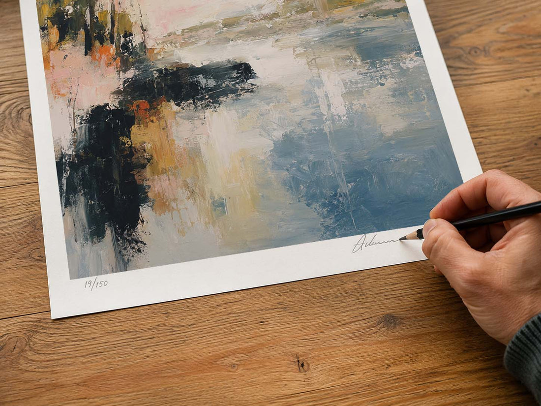 A hand holding a pencil signs a limited edition abstract art print on a wooden table. The print features a white border and an abstract painting with blue, gray, black, beige, and orange tones.