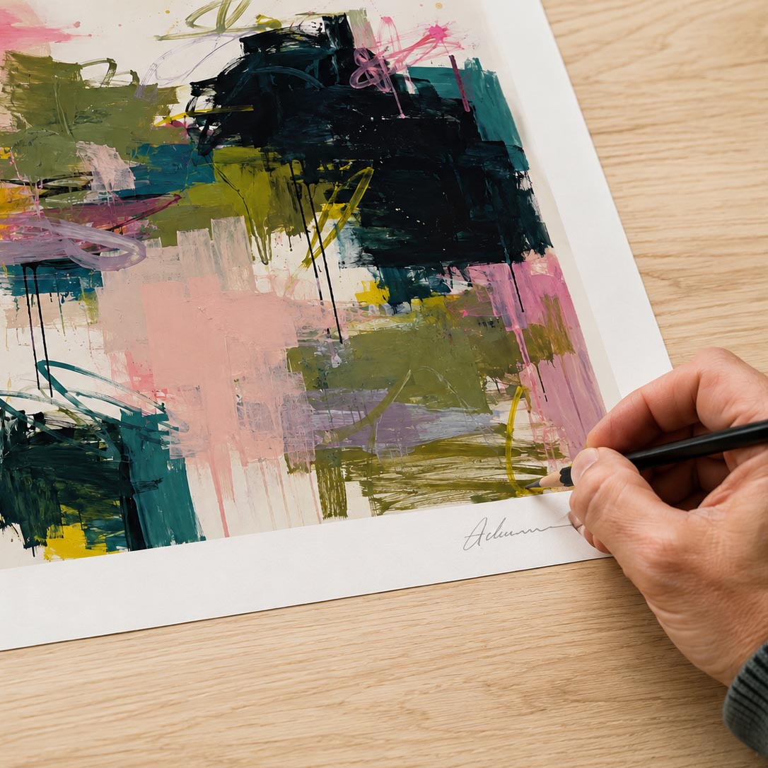 An abstract art print with colorful brushstrokes in green, pink, black, and yellow, shown on a light wood table with a hand holding a pencil near the bottom right corner.
